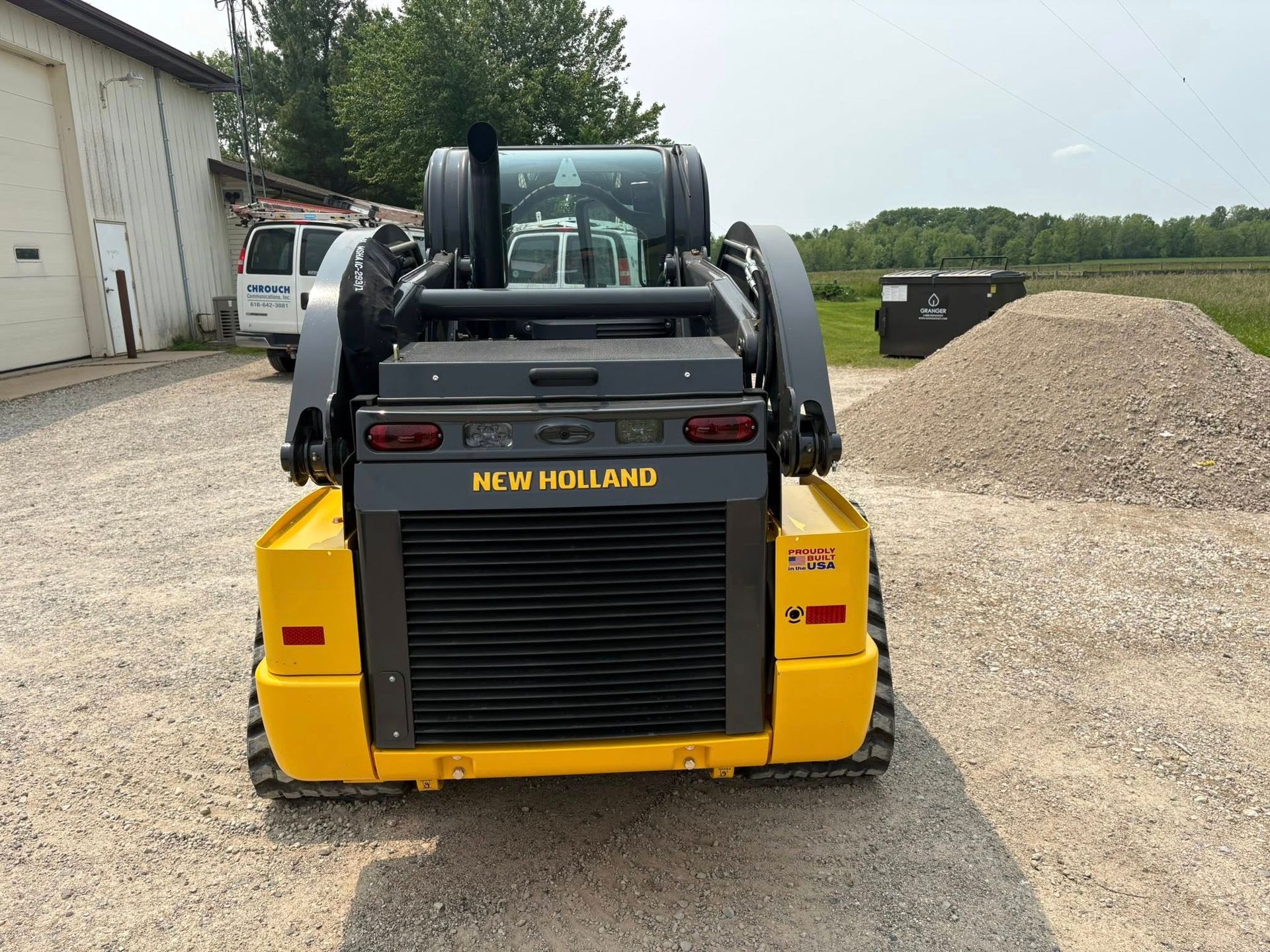 Yellow New Holland skid steer loader parked on gravel beside a building and gravel pile