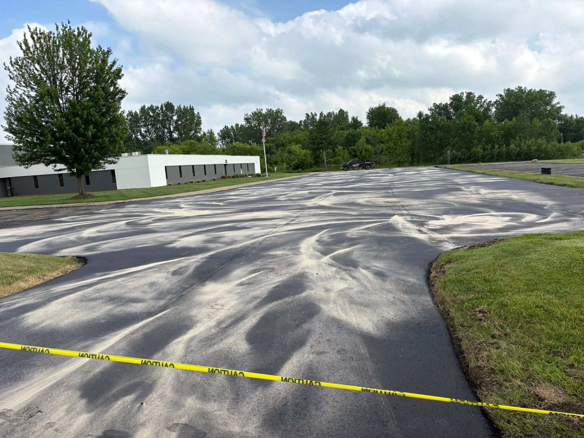 Empty paved lot with yellow caution tape, trees, and a white building under a cloudy sky