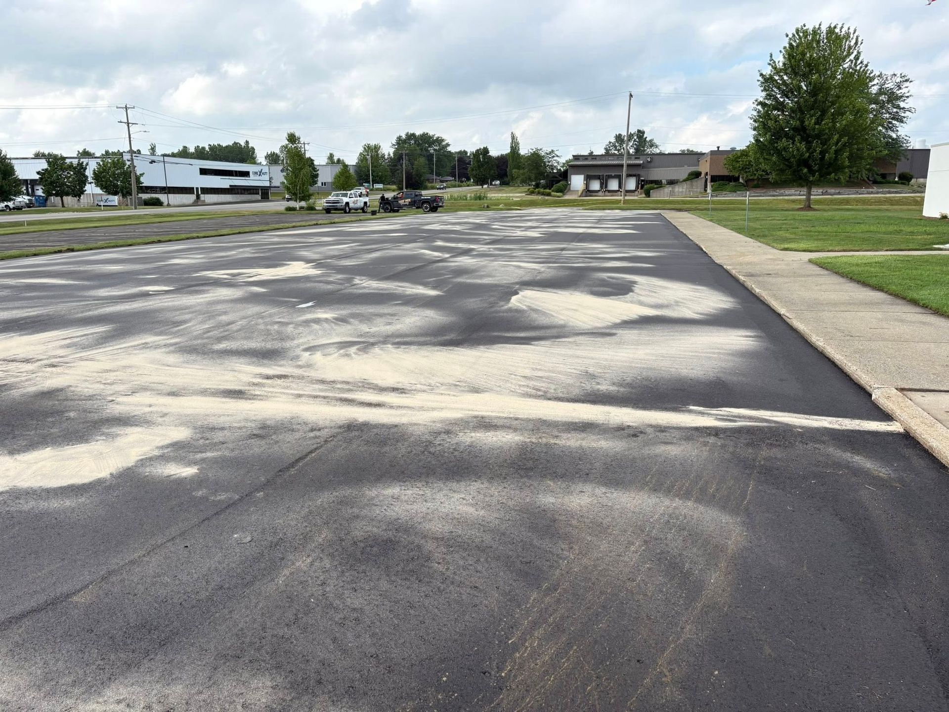 Empty asphalt parking lot with cracked patches, a sidewalk, and trees under a cloudy sky
