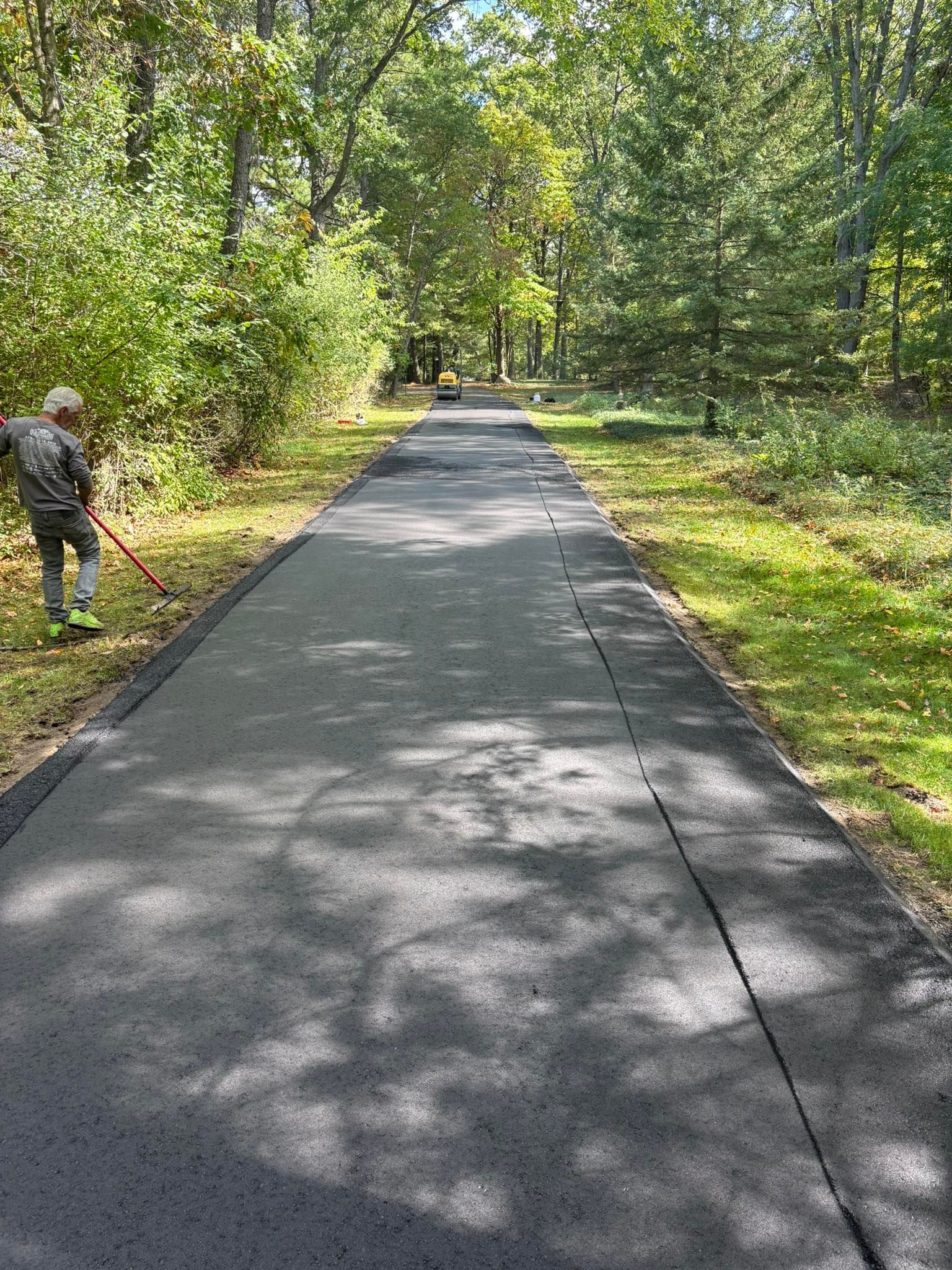 A paved path through a wooded park, with a person working along the left side using a tool.