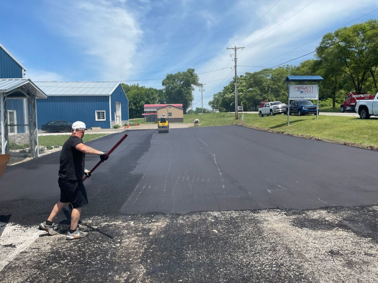 Person spreading fresh blacktop on a paved road near blue buildings on a sunny day