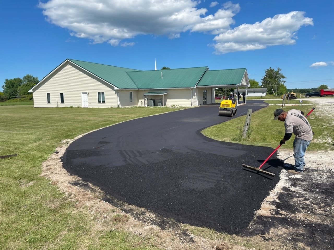 Worker paving a curved driveway beside a green-roofed building under a blue sky