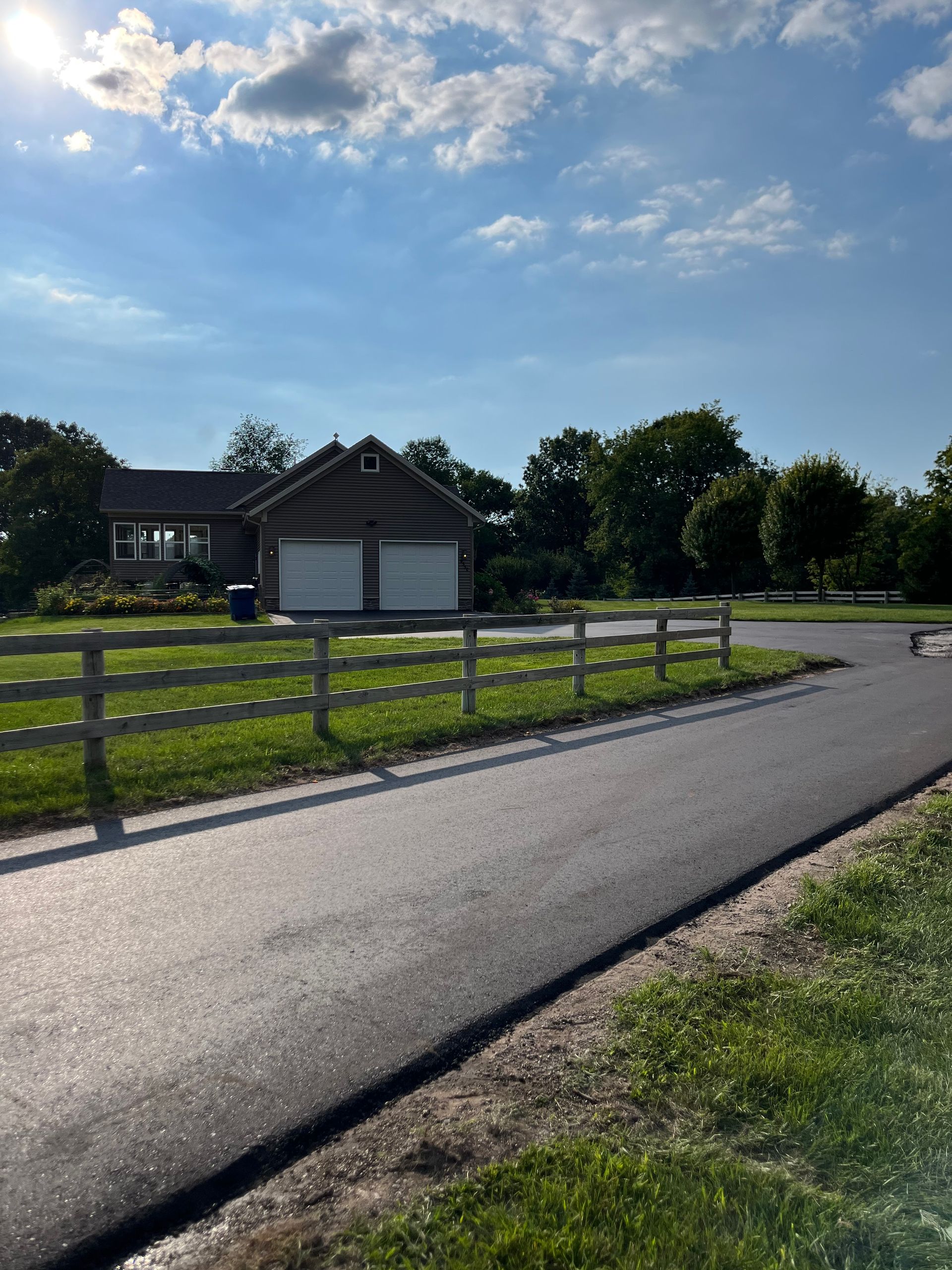 Rural road beside a fenced green field and small barn under a bright blue sky