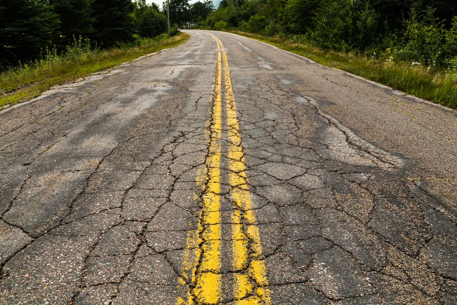 Cracked rural road with faded double yellow lines, bordered by trees and forest greenery