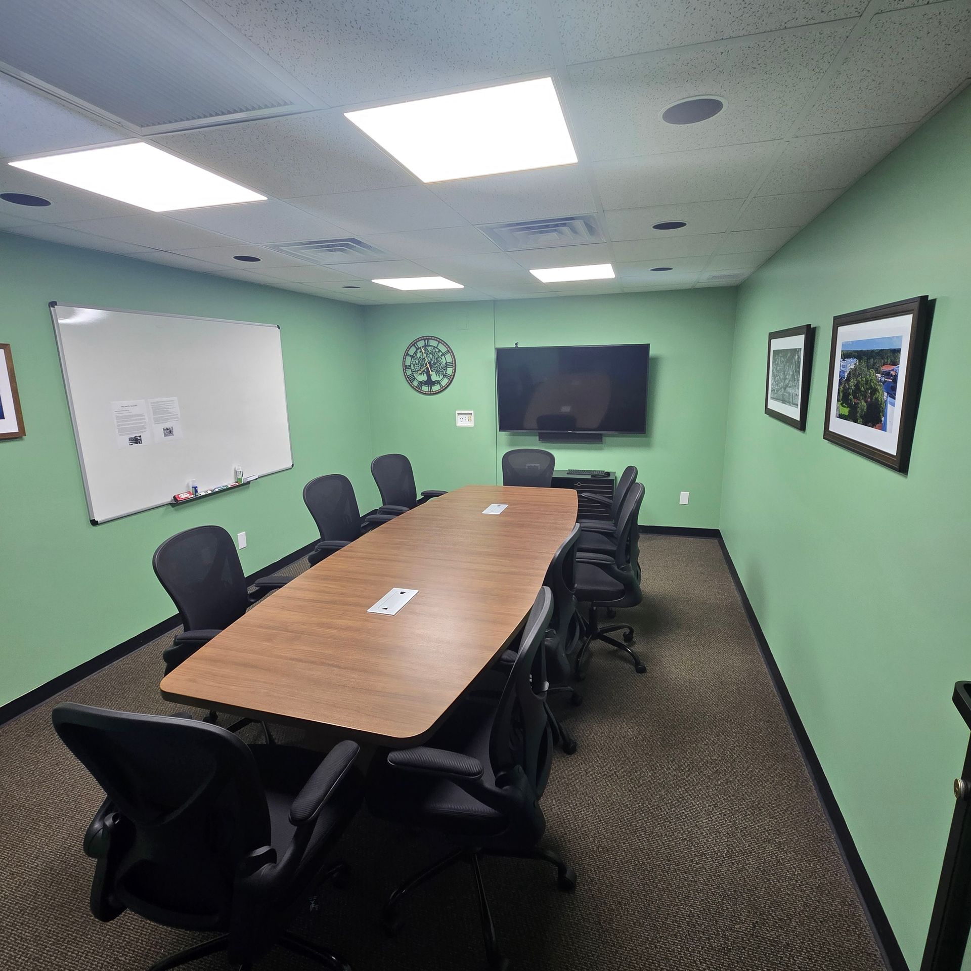 Conference room with long table, black chairs, whiteboard, wall-mounted TV, and green walls.