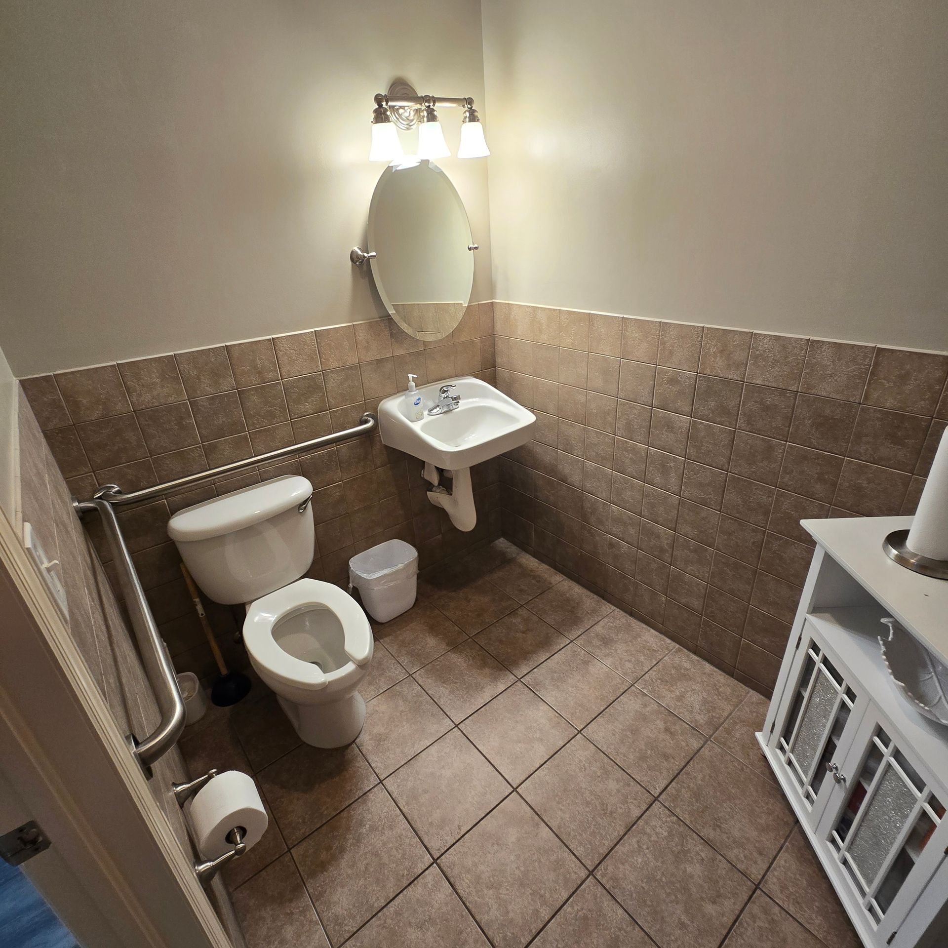 Small bathroom with toilet, sink, mirror, tiled walls and floor, and a shower hose on the left.