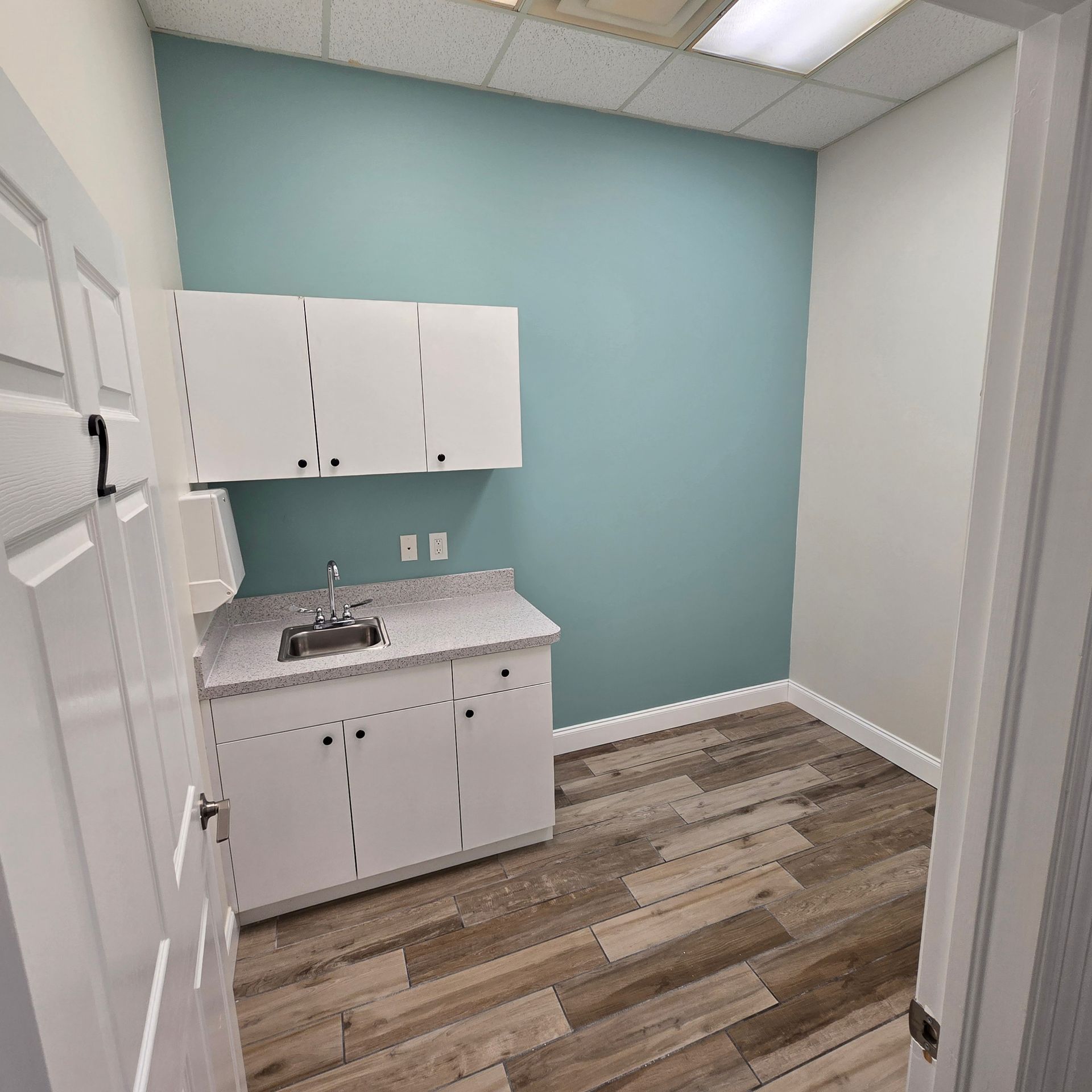 Small empty break room with white cabinets, sink, and teal walls under fluorescent lighting.