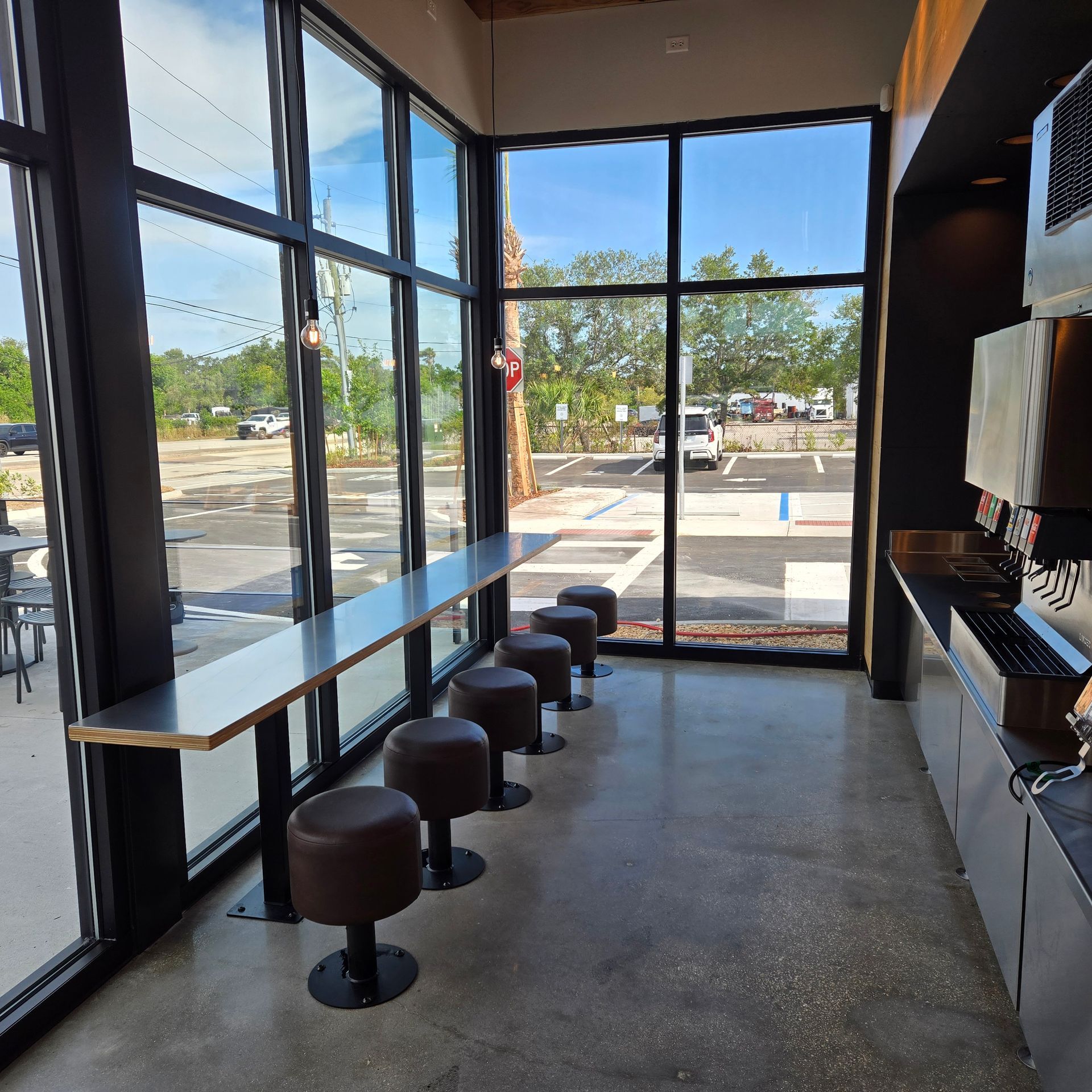 Empty cafe seating area with stools by large windows overlooking a bright street.