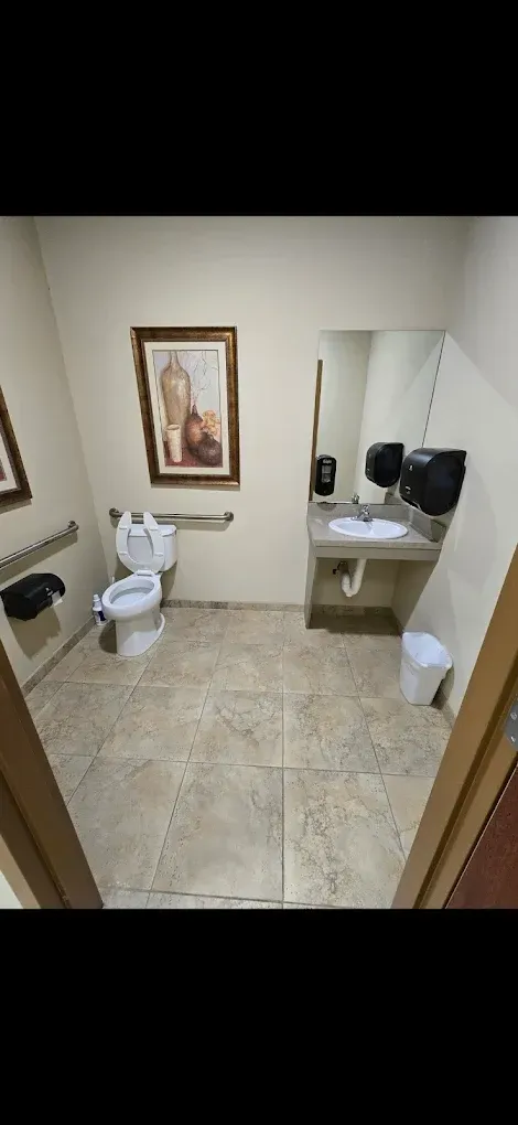 Small beige bathroom with toilet, sink, mirror, framed art, and a wall-mounted paper towel dispenser.