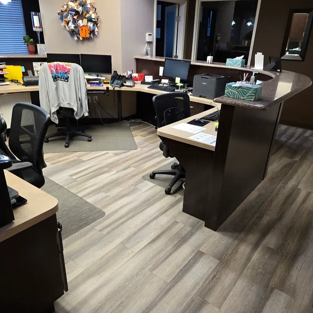 Reception desk in office. Light wood floors and countertops. Desks with computers and chairs.