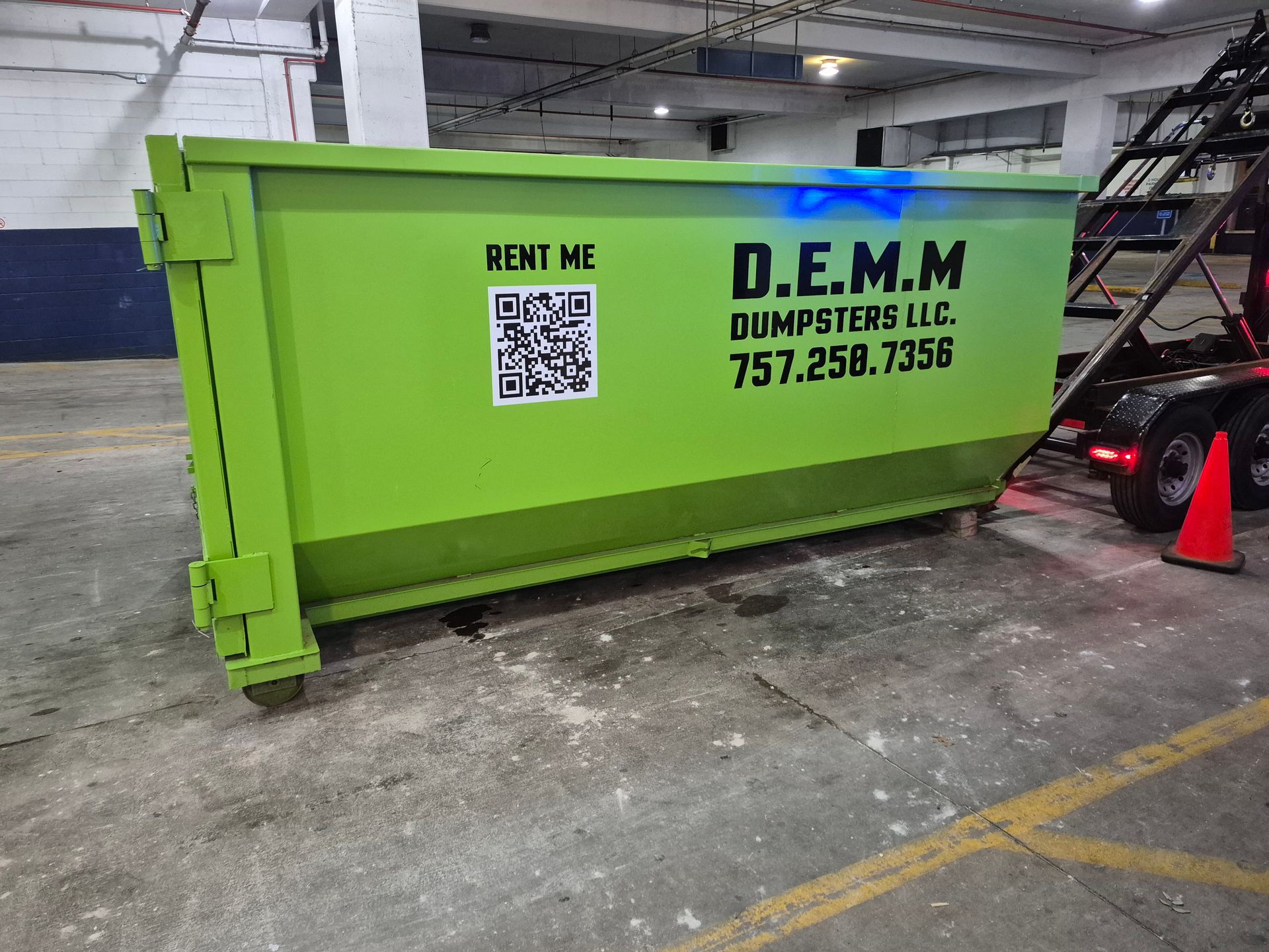 Bright green dumpster on a black trailer, outdoors. A long metal rod supports the lid.