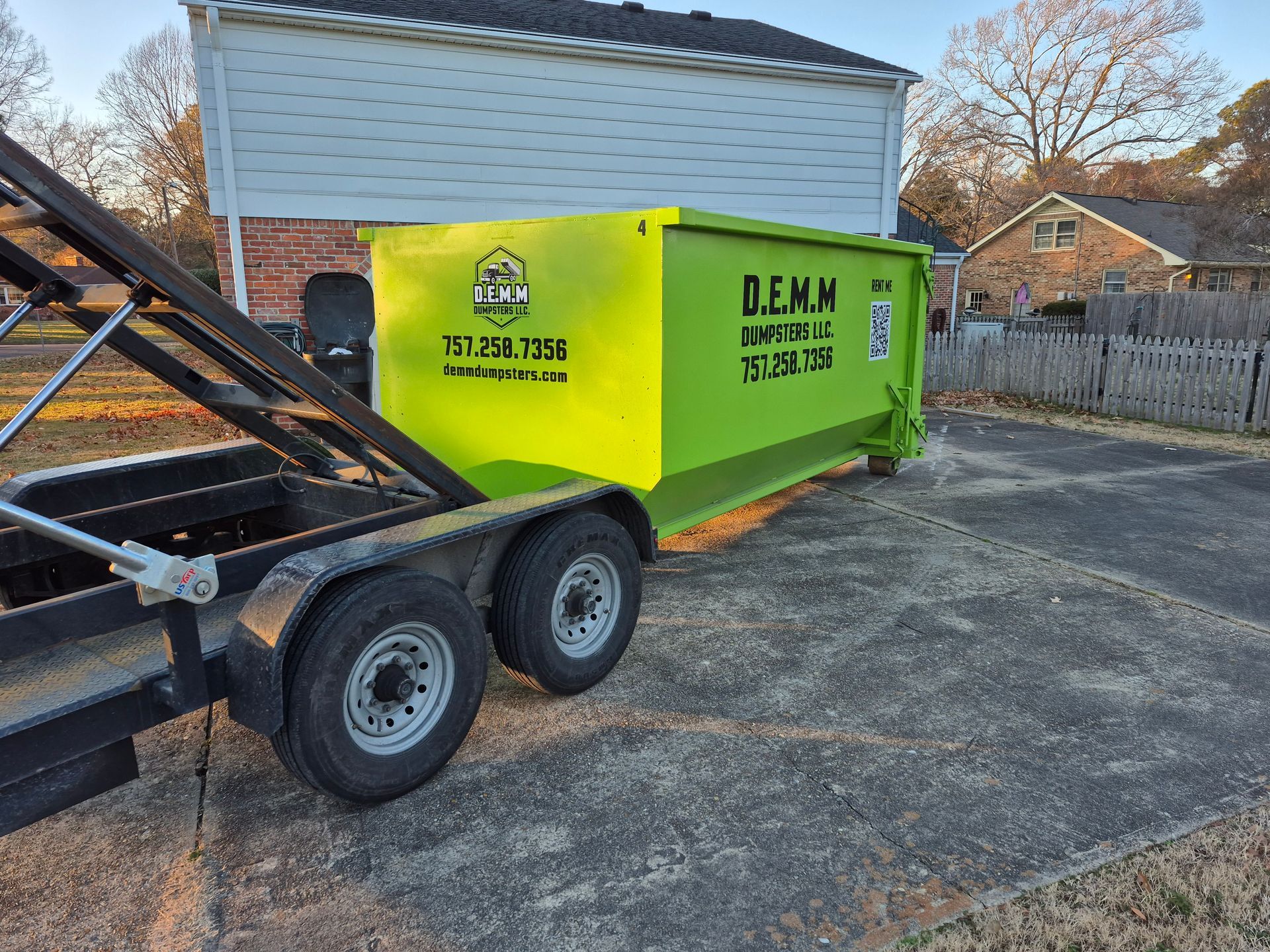 A blue pickup truck pulling a black dumpster trailer parked next to a house with a white garage.