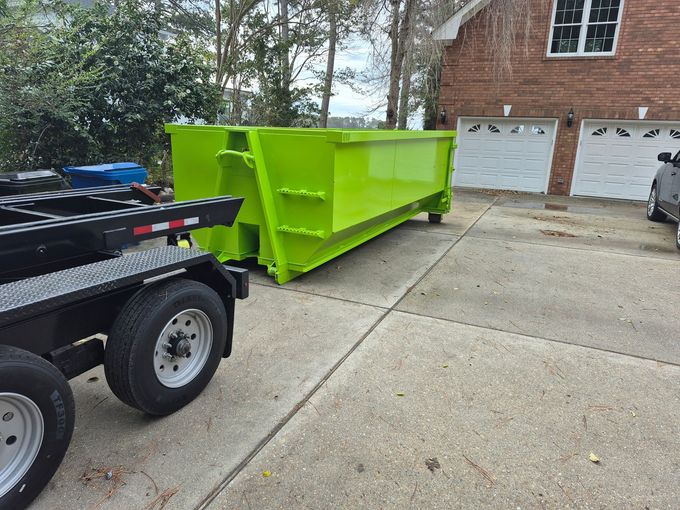 A blue truck hauling a large black dumpster trailer parked on a driveway next to a house with a white garage door.