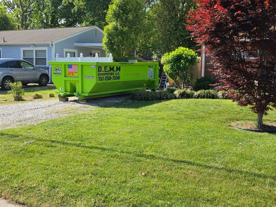 A blue pickup truck with a large black dumpster trailer parked in a driveway next to a brick house with a white garage door.