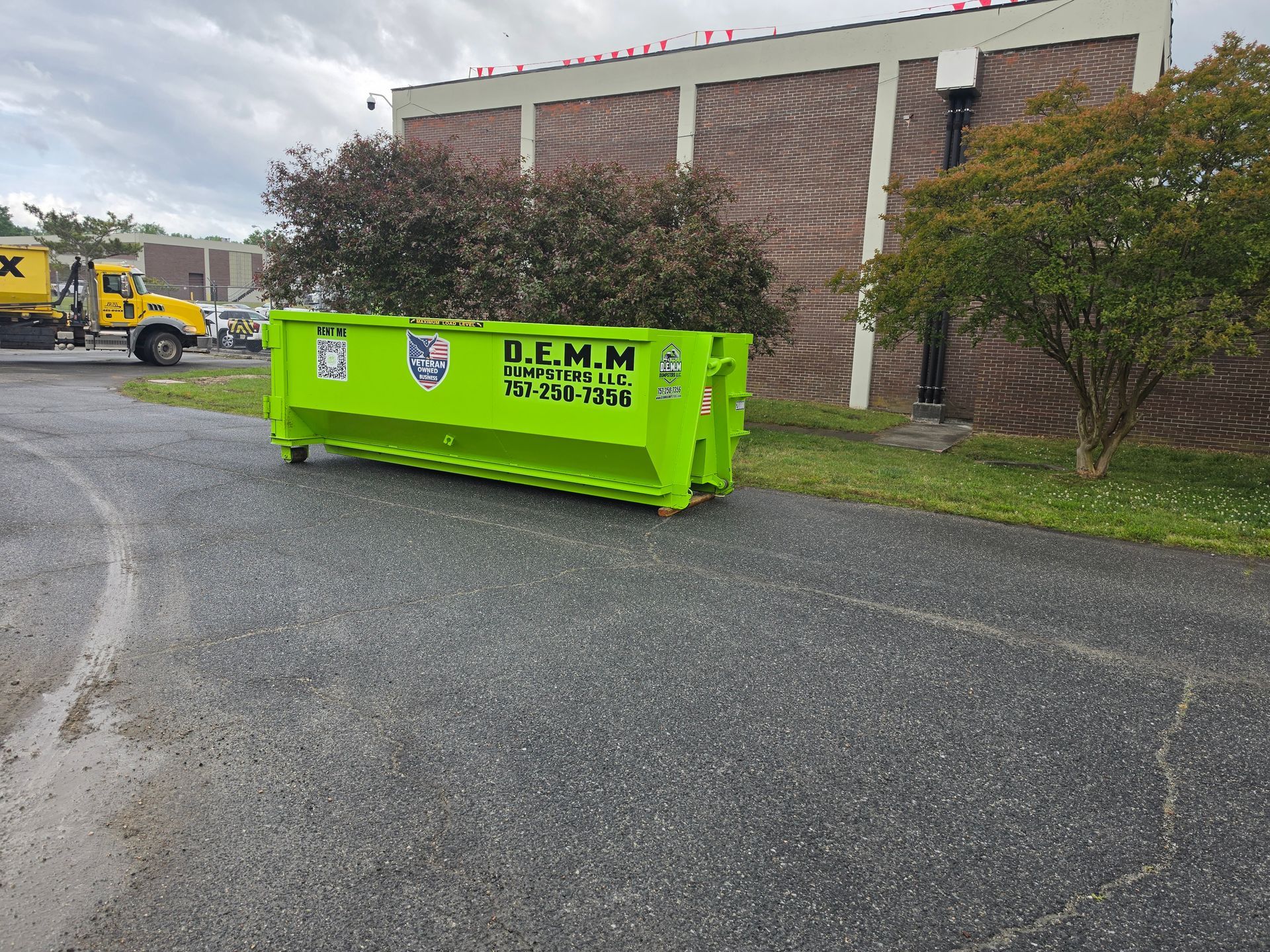 A bright green dumpster next to a yellow forklift on a concrete surface, with a trailer and a building in the background.