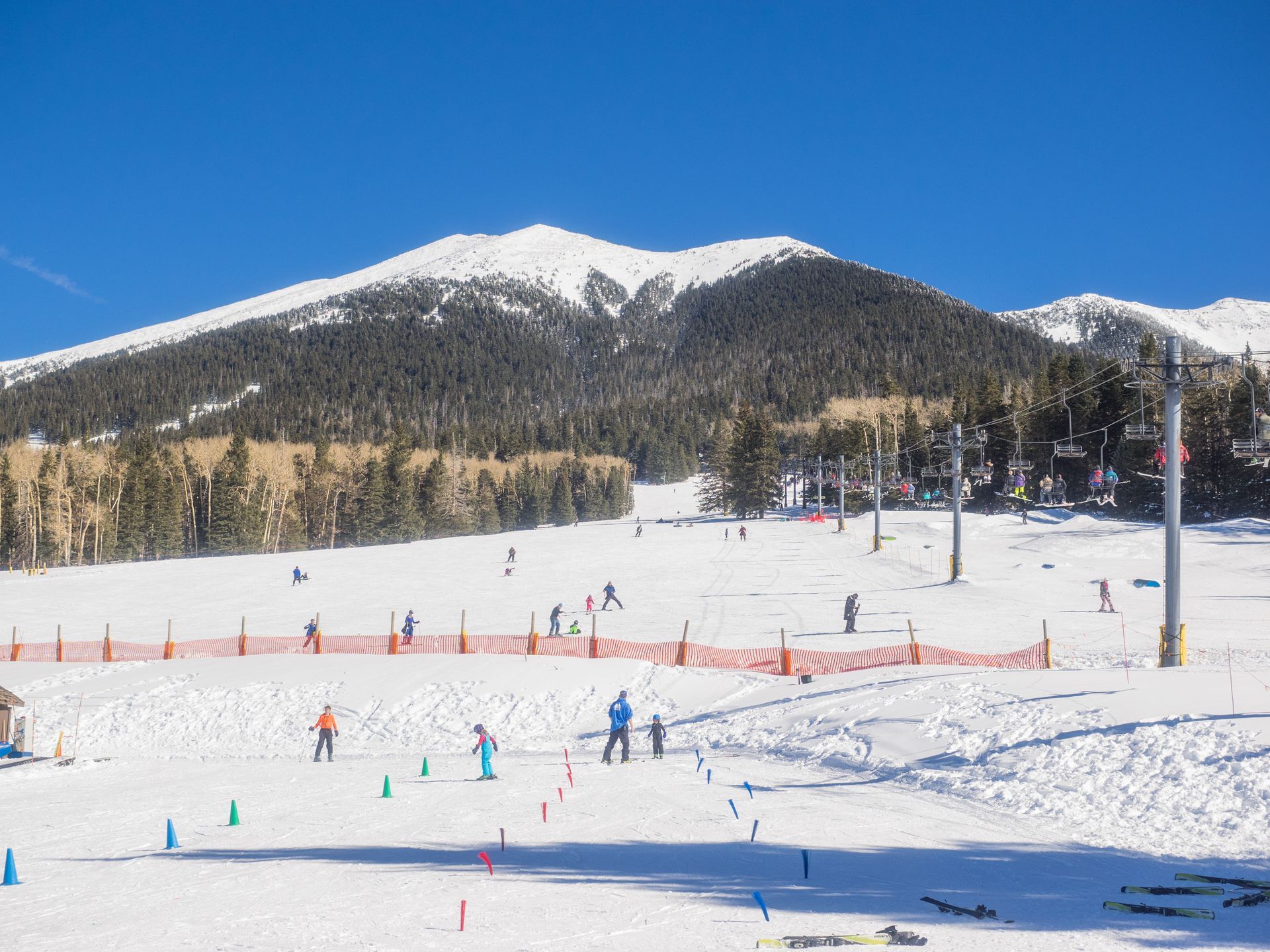 Snowy ski slope with skiers, orange fencing, and a forested mountain under a blue sky