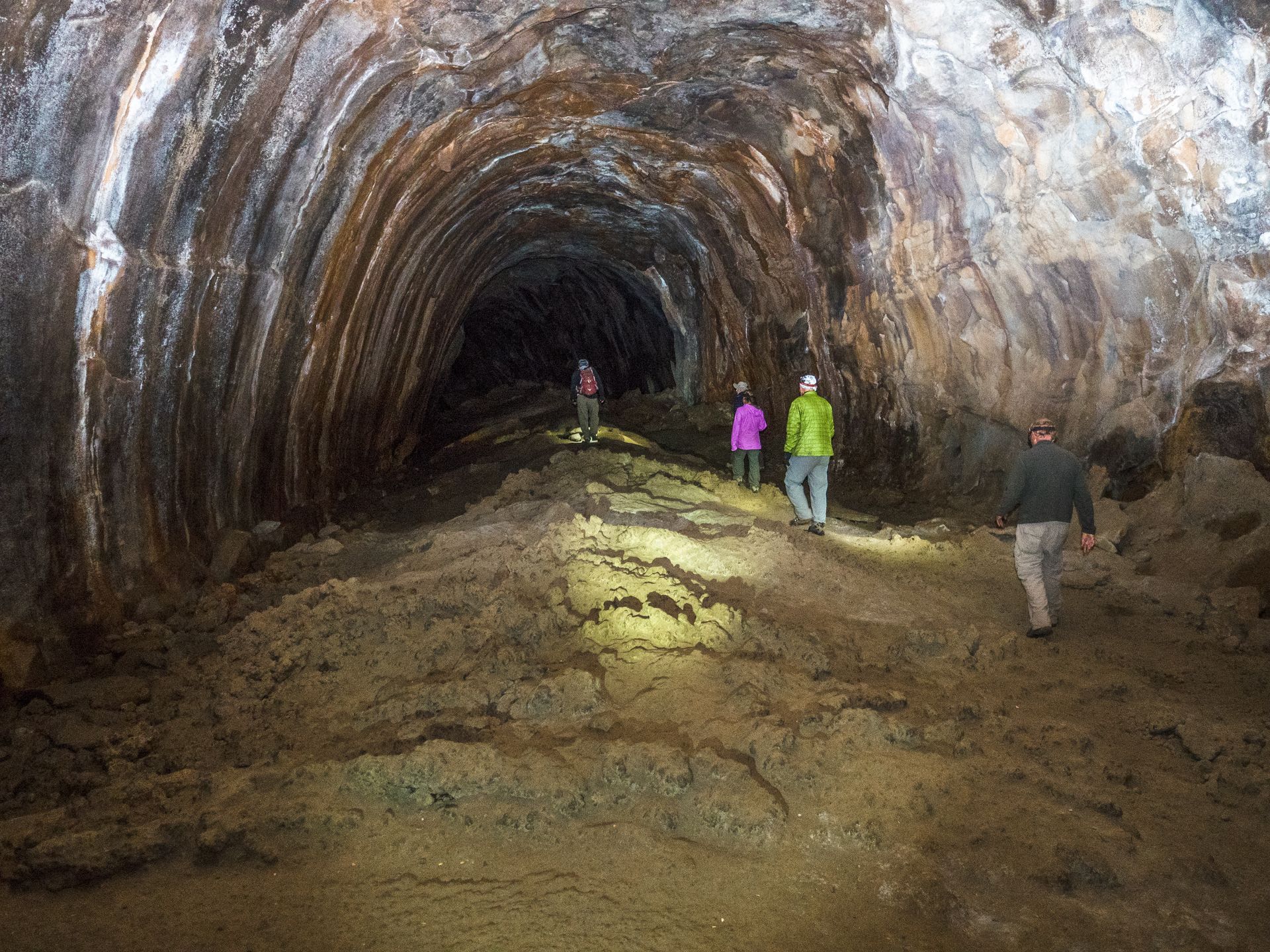 People walking through a dimly lit cave tunnel with rocky walls and muddy ground.
