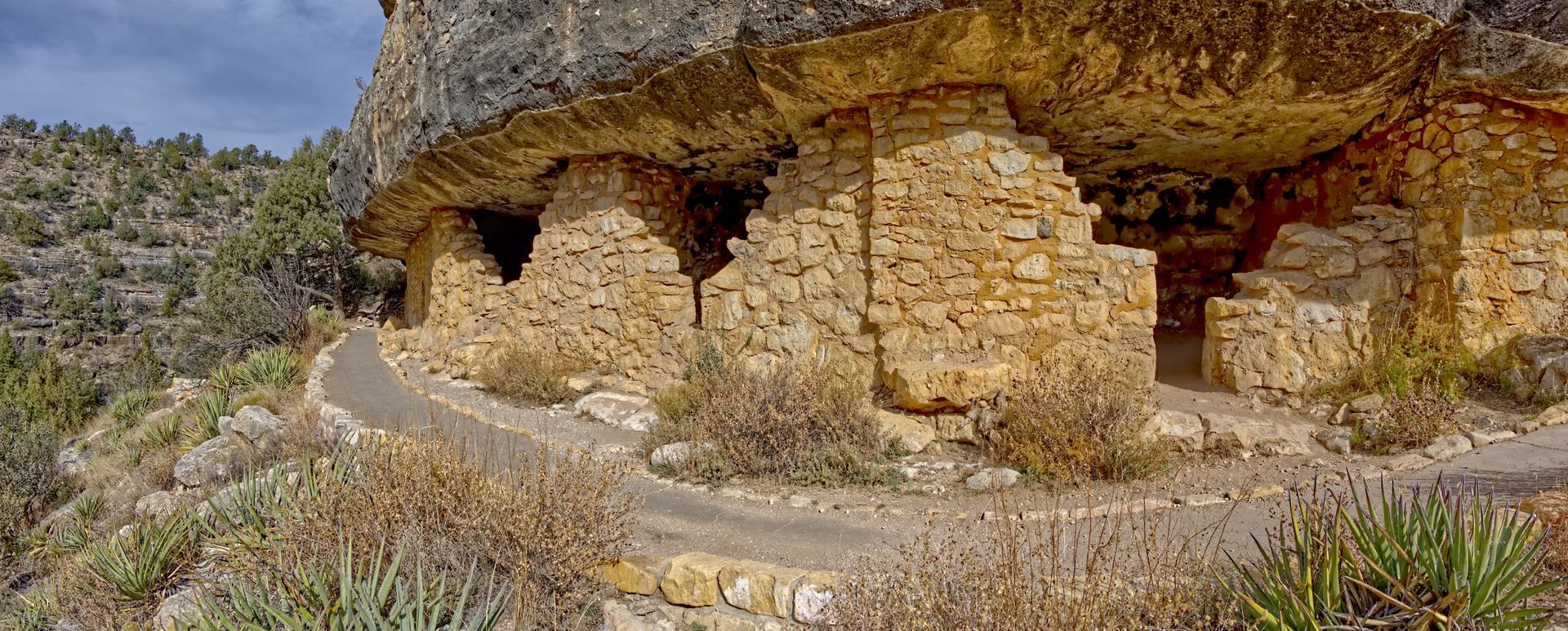 Stone cliff dwellings carved into a rocky hillside beside a winding path in a dry landscape