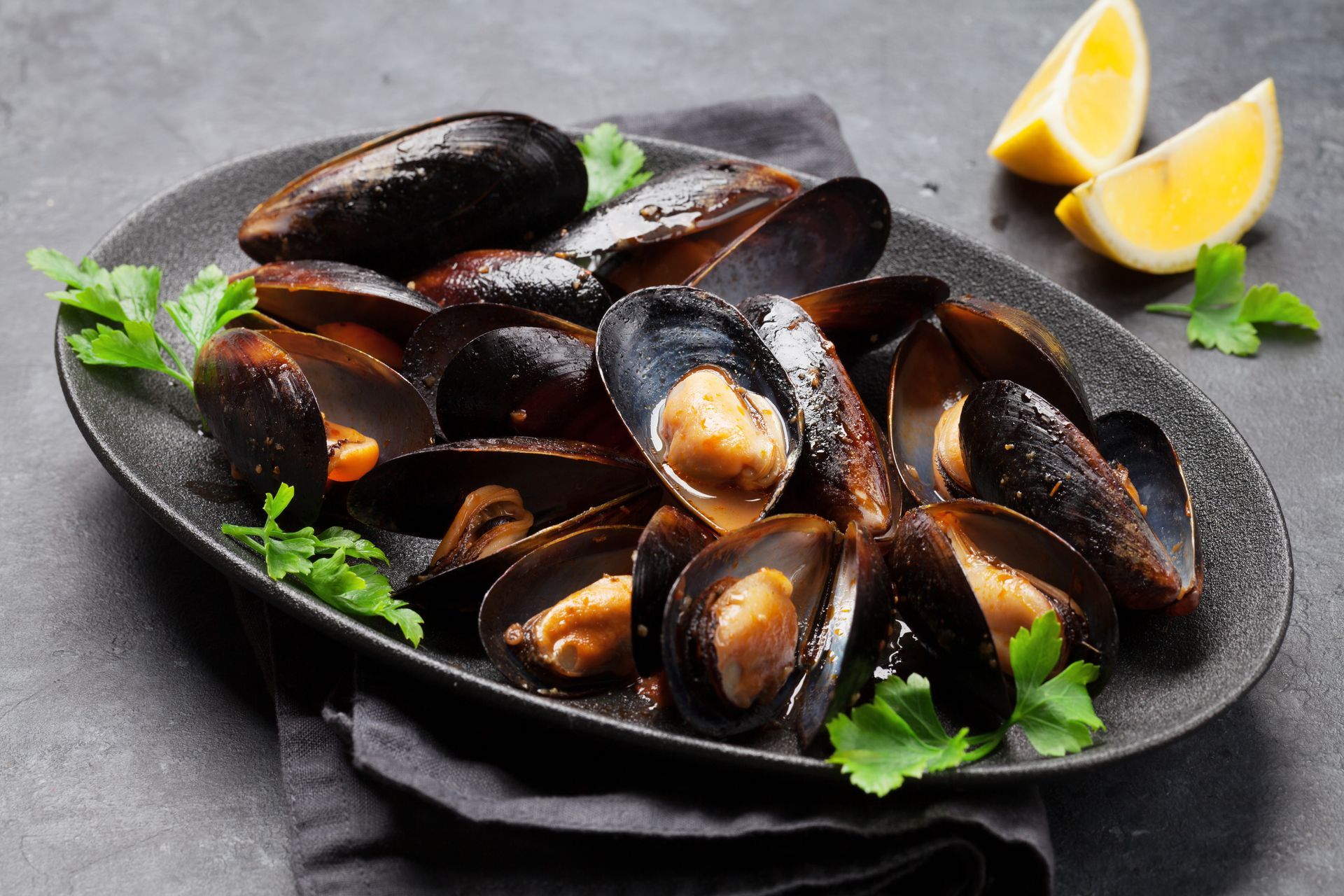 Plate of steamed mussels with lemon wedges and herbs on a dark slate surface