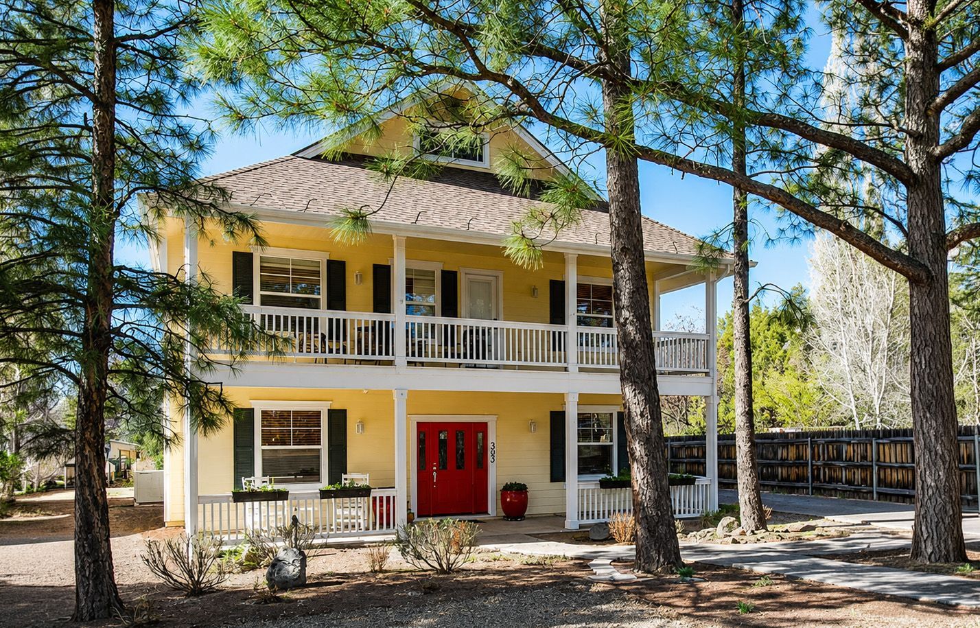 Two-story yellow house with white porch and red front door among tall pine trees