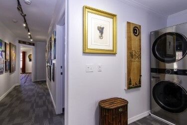 Laundry hallway with framed wall art, a wooden stool, and stacked washer and dryer units.
