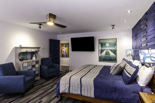 Modern bedroom with a blue bed, armchairs, wall art, and a TV on a white ceiling with a fan