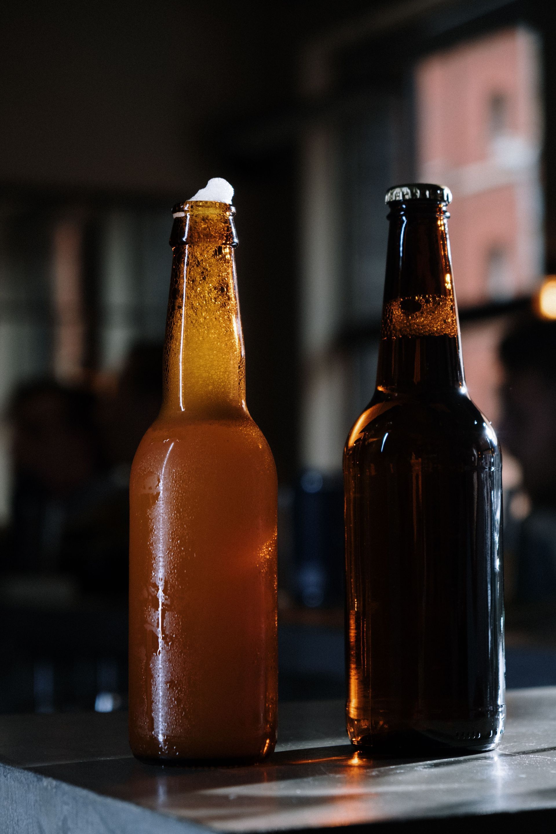 Two beer bottles on a table, one amber and one dark, against a blurred indoor background