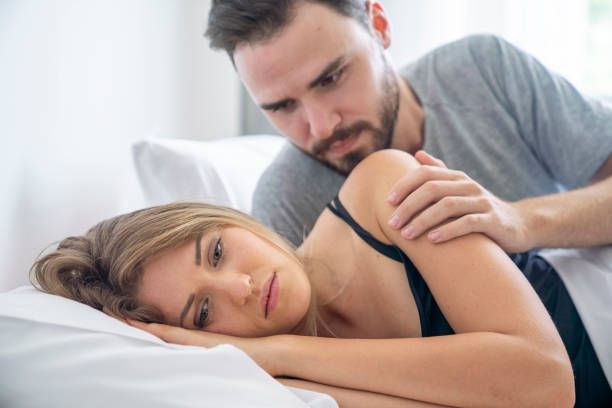 A man comforts a woman lying in bed, hand on her shoulder, both with concerned expressions.