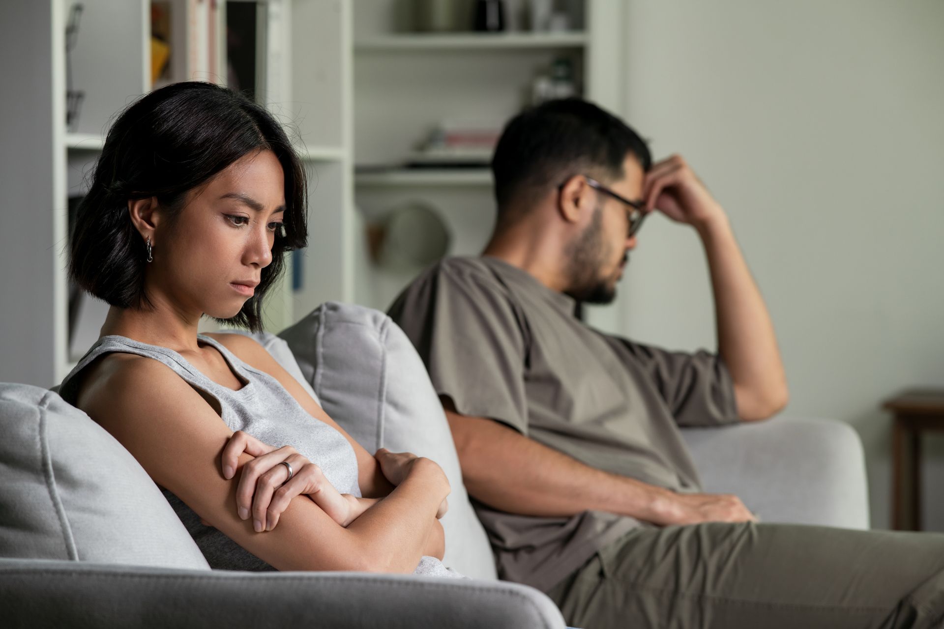 Woman with arms crossed, man with hand on forehead, both looking sad on a couch.