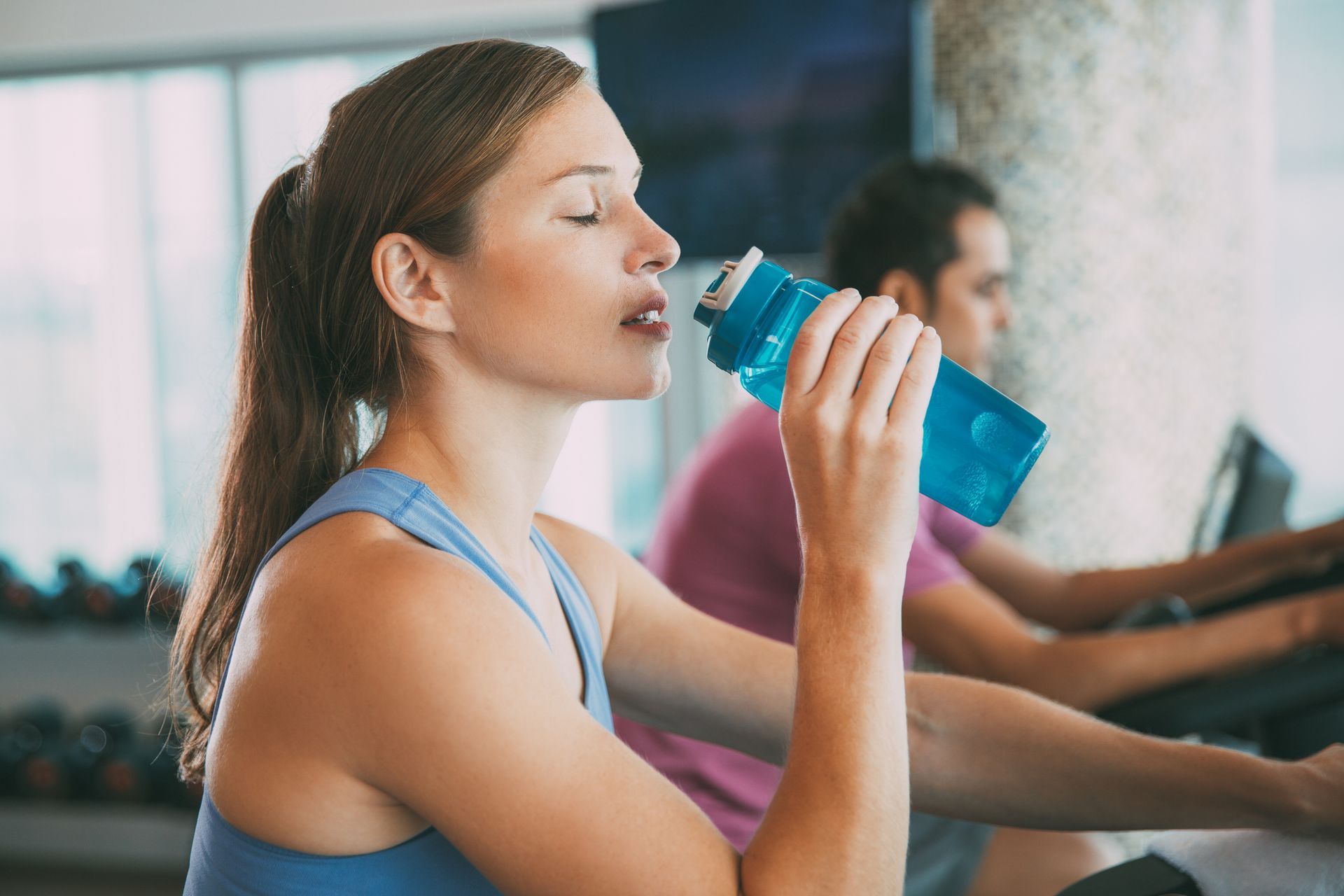 Person hydrating with blue water bottle while exercising on stationary bike in modern gym.