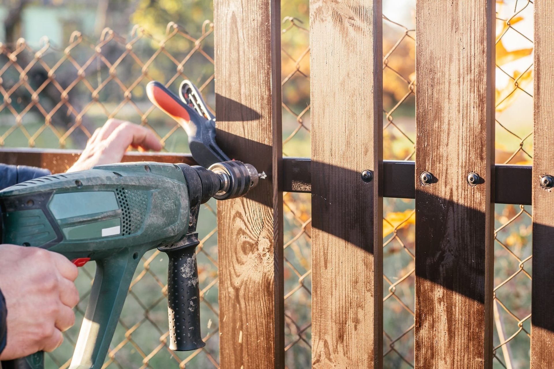 A person uses a power drill to fasten a wooden board onto a metal fence frame outdoors.