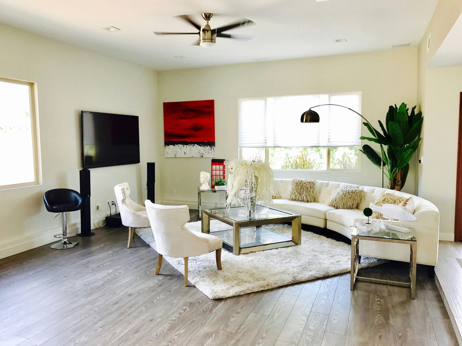 A modern living room with a white curved sofa, two white chairs, a glass coffee table, and large windows on a rug.