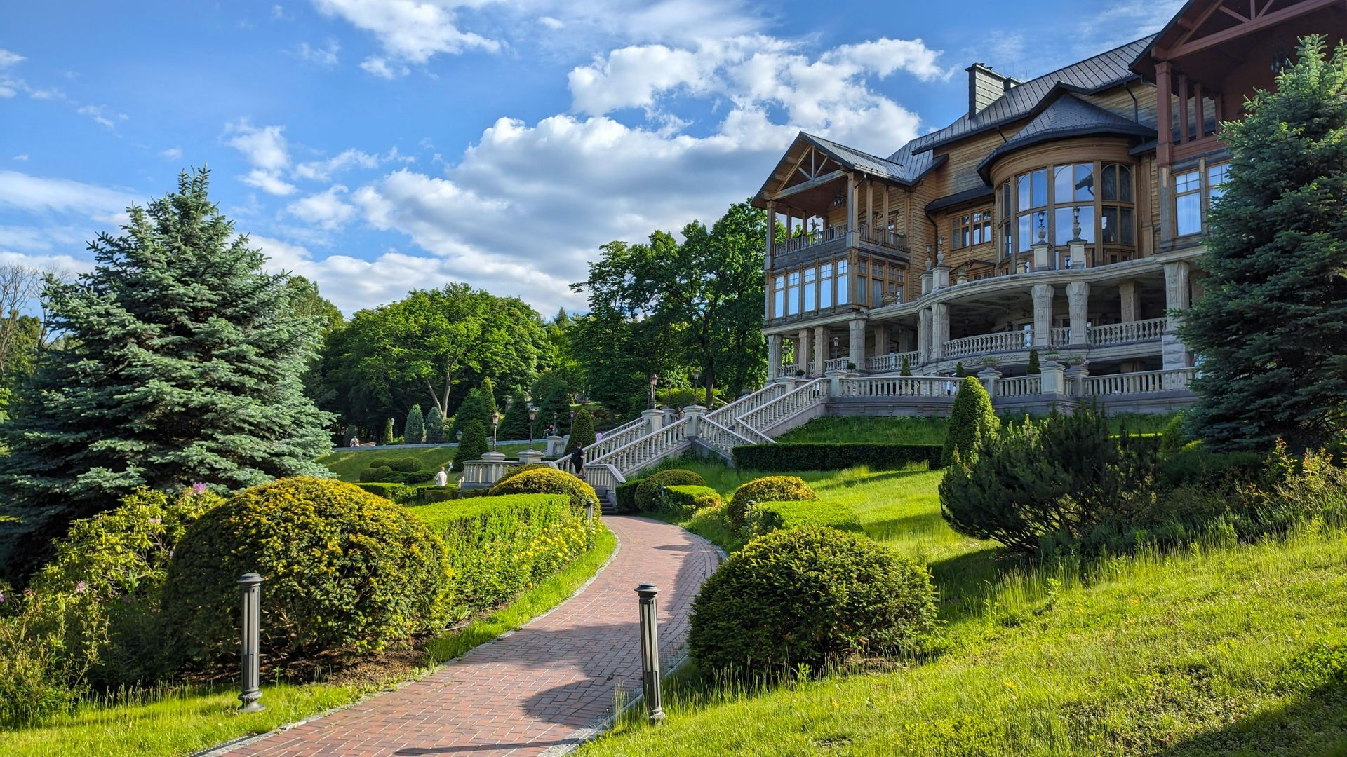 A stone path winds through a green garden toward a large wooden mansion with a stone staircase under a blue, cloudy sky.