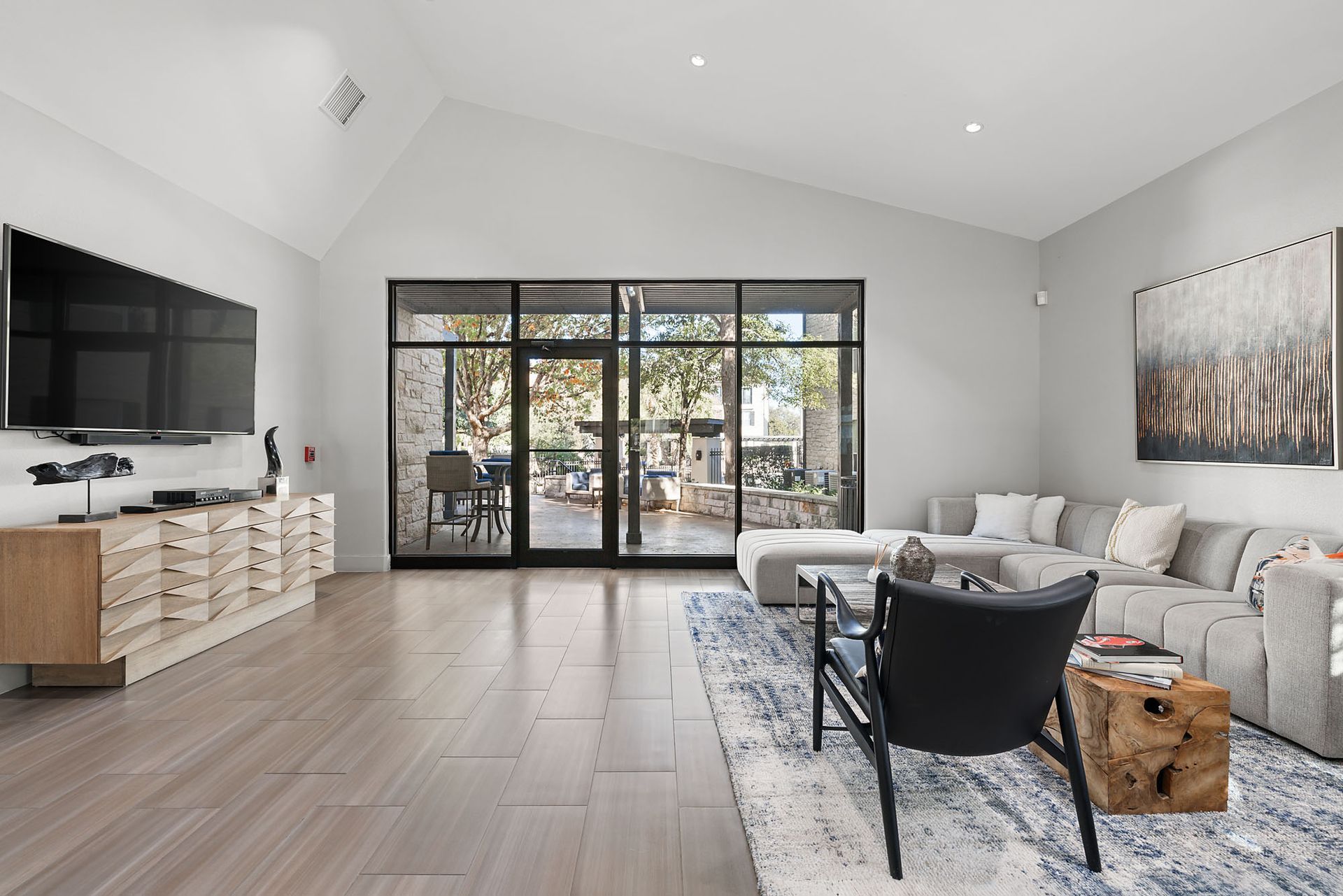 Bright modern living room with gray sofa, black chair, TV, and glass doors to a patio at Marquis at Great Hills in Austin, TX.