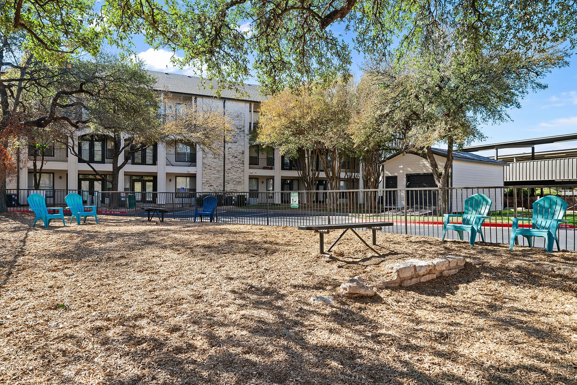 Apartment courtyard with turquoise chairs, picnic tables, and trees in front of a building at Marquis at Great Hills in Austin, TX.