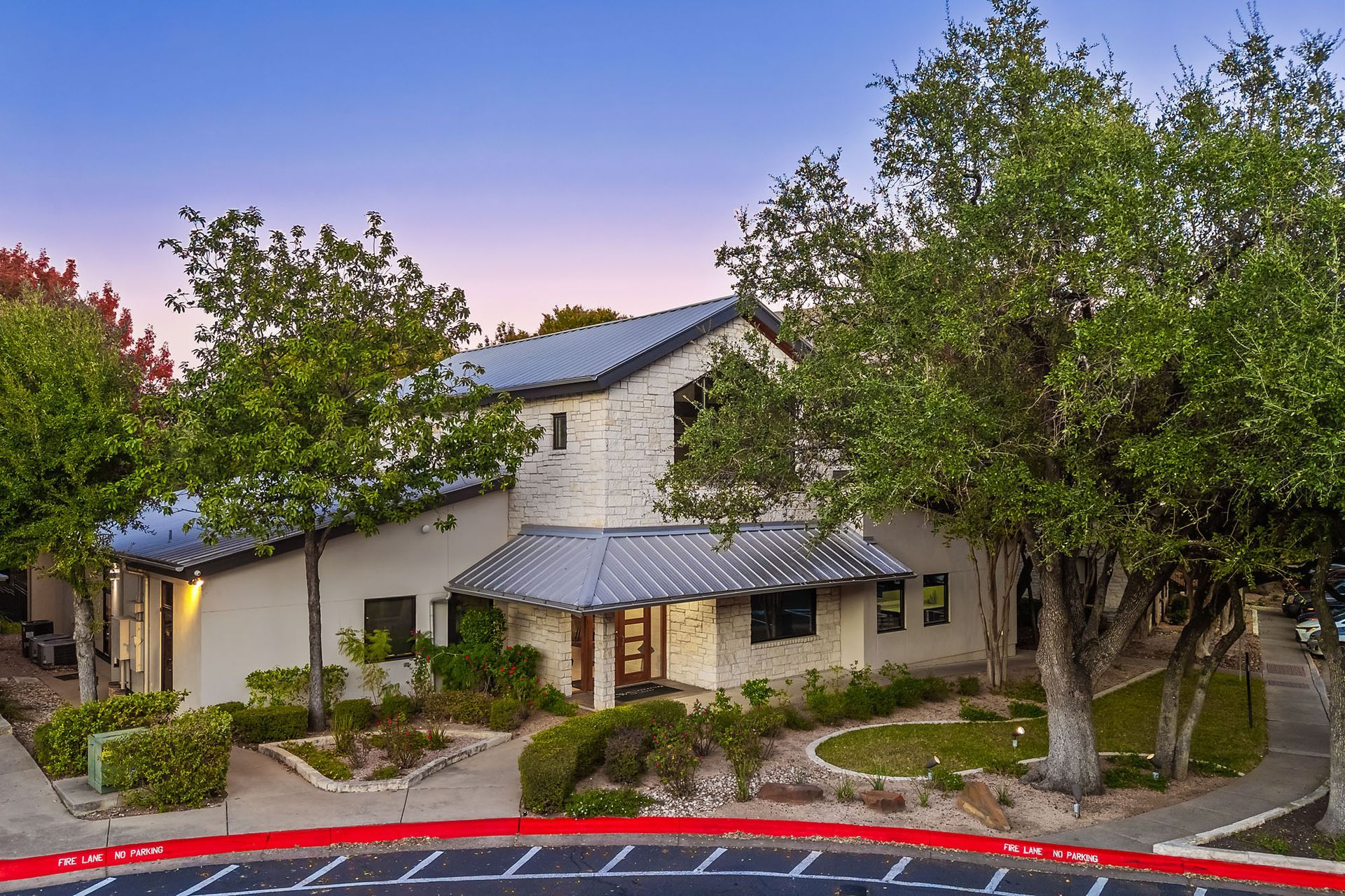 Two-story beige building with trees, shrubs, and a red curb at dusk at Marquis at Great Hills in Austin, TX.