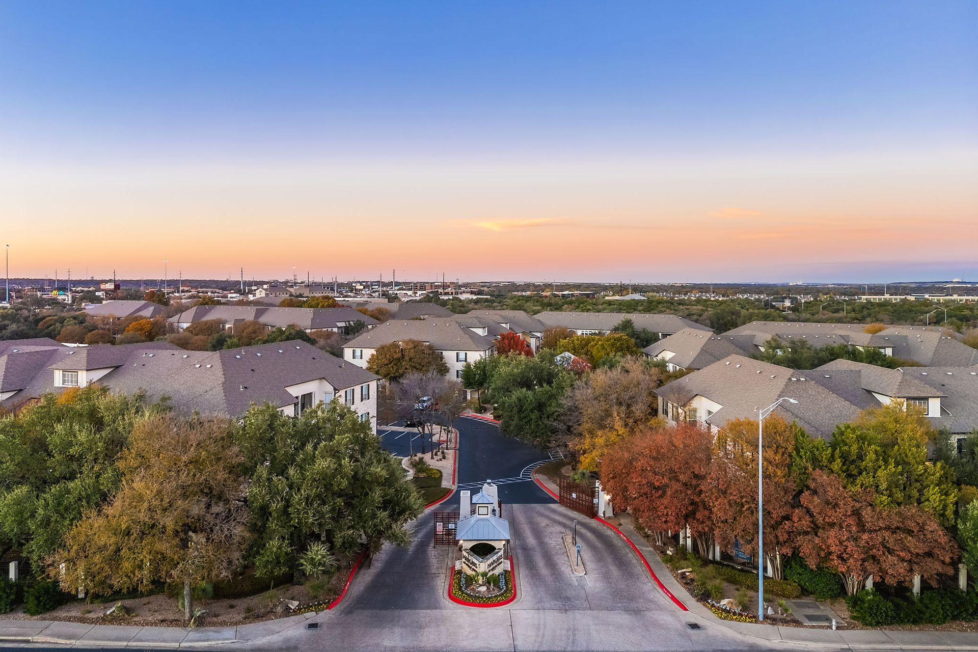 Sunset view over a suburban neighborhood entrance with houses, trees, and a central monument island at Marquis at Great Hills in Austin, TX.
