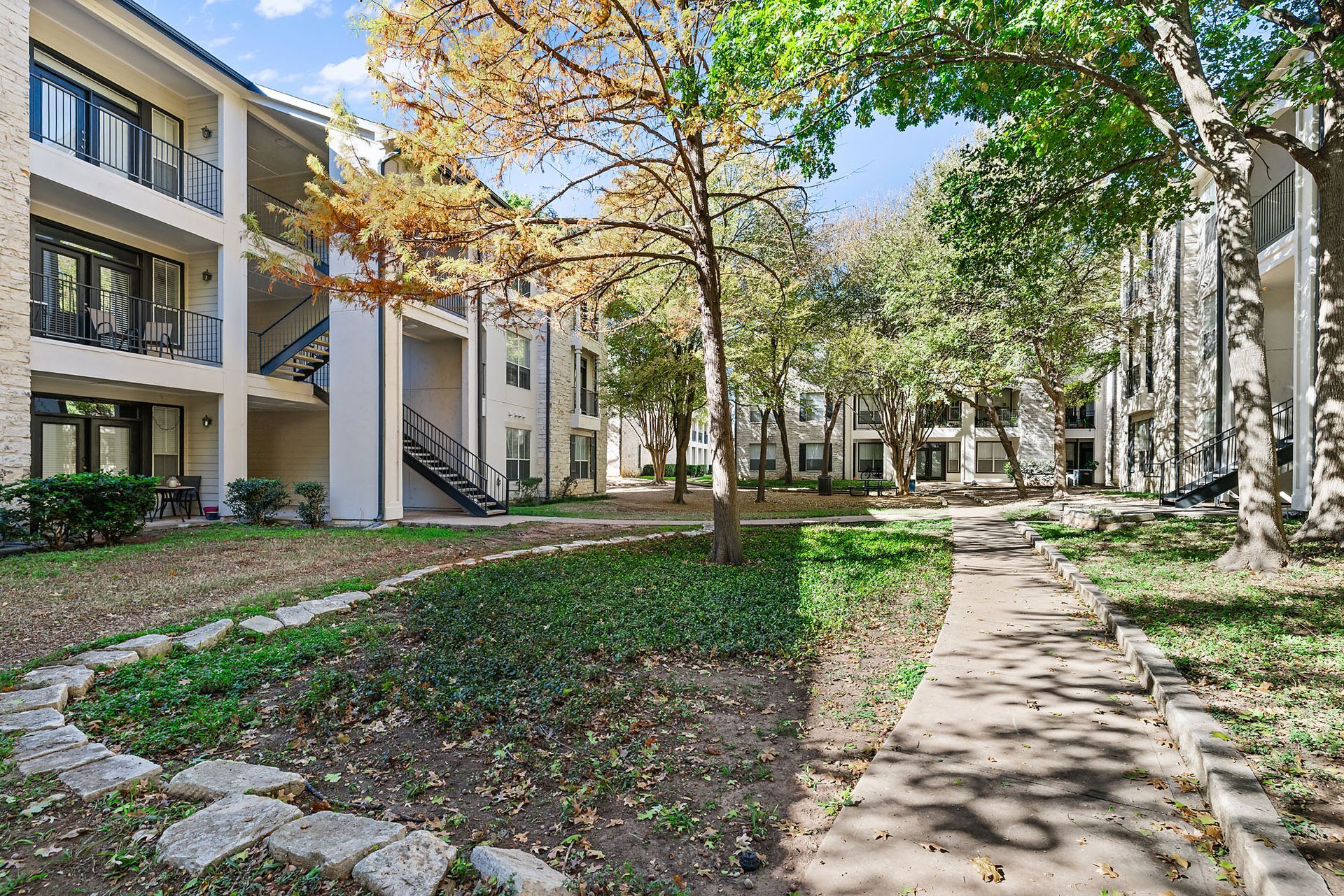 Apartment courtyard with trees, grass, and a stone walkway beside beige buildings at Marquis at Great Hills in Austin, TX.