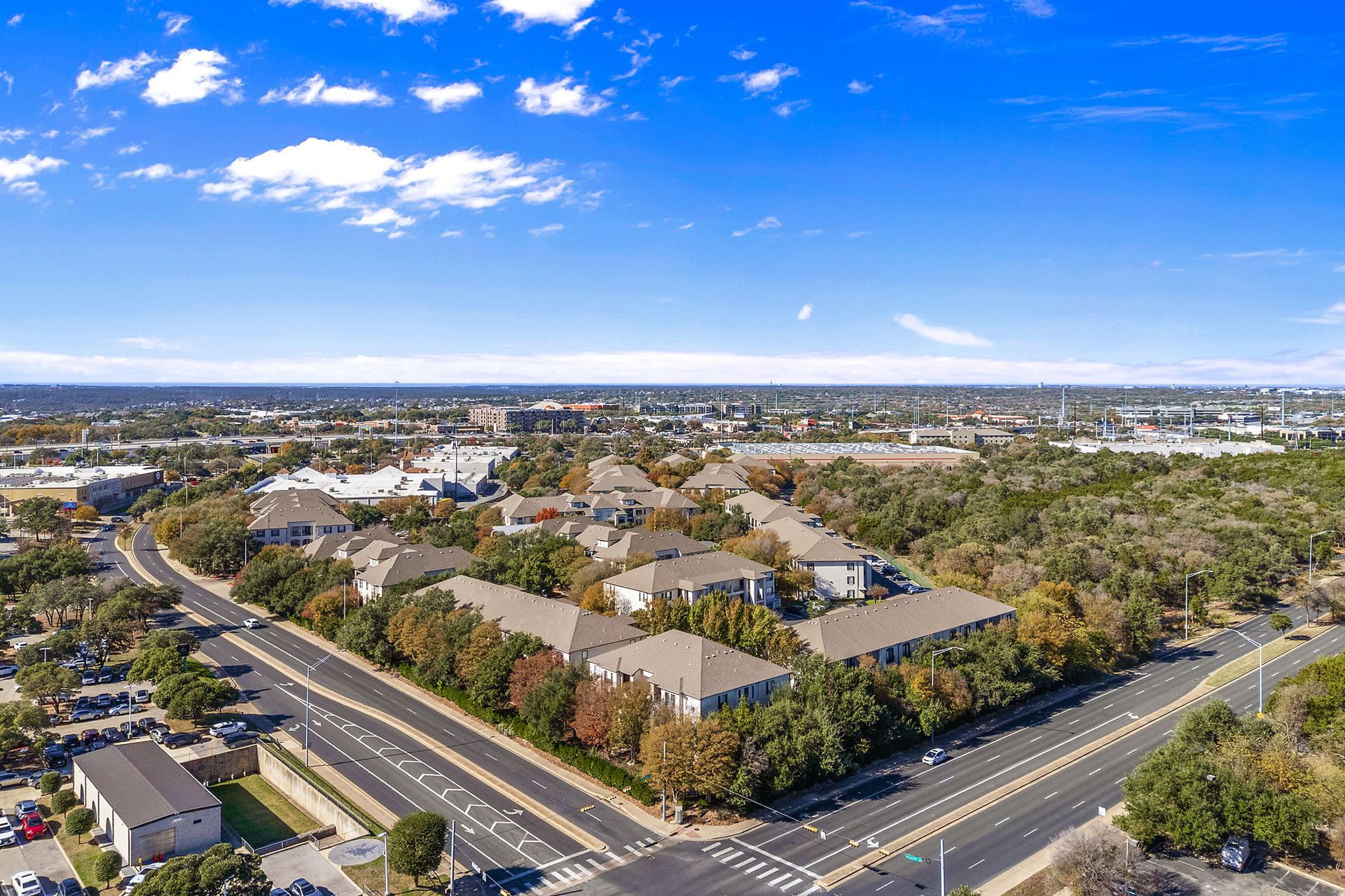 Aerial view of a suburban neighborhood with roads, buildings, and trees under a bright blue sky at Marquis at Great Hills in Austin, TX.