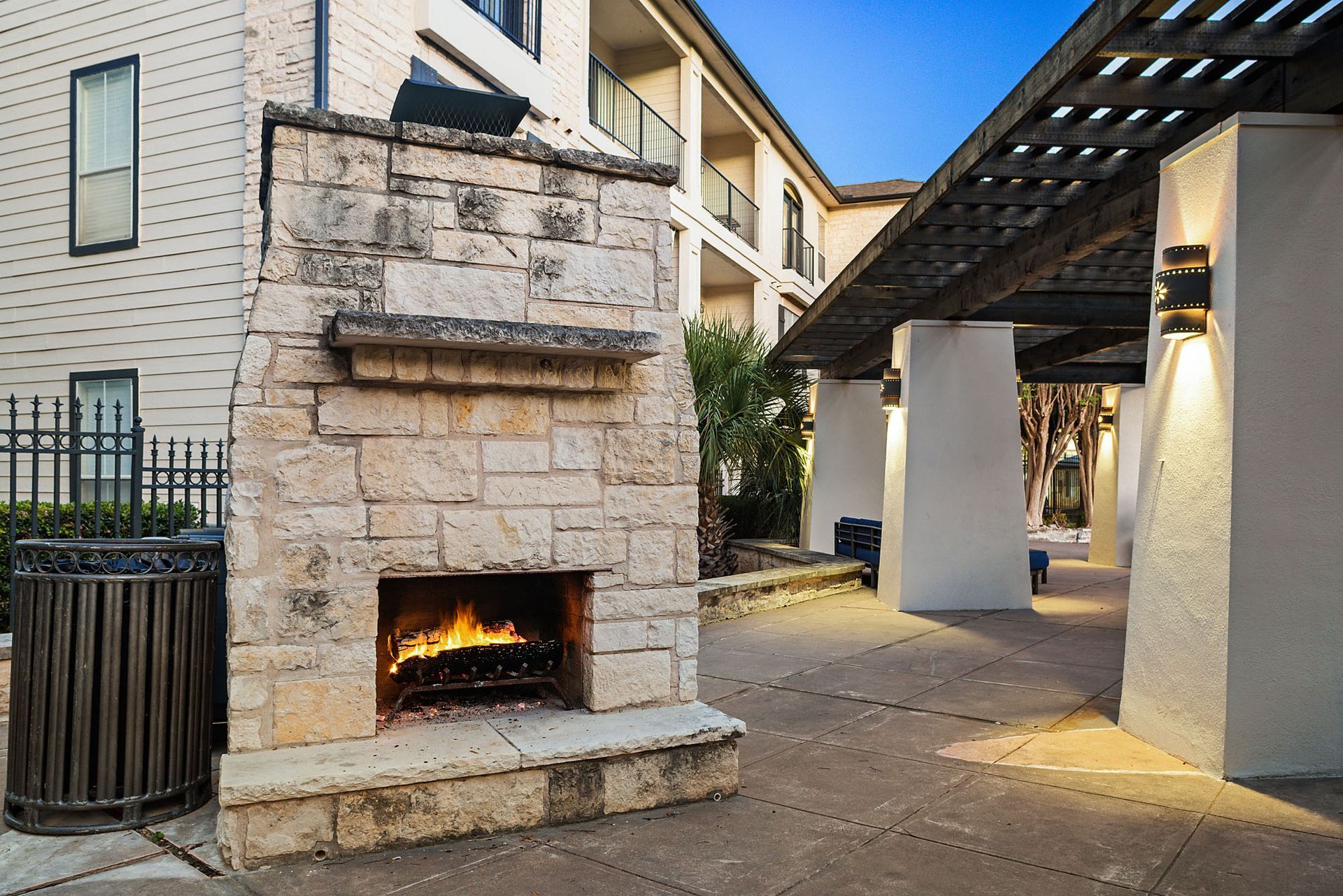 Stone outdoor fireplace with a lit fire in a courtyard beside a building and pergola-covered walkway at Marquis at Great Hills in Austin, TX.
