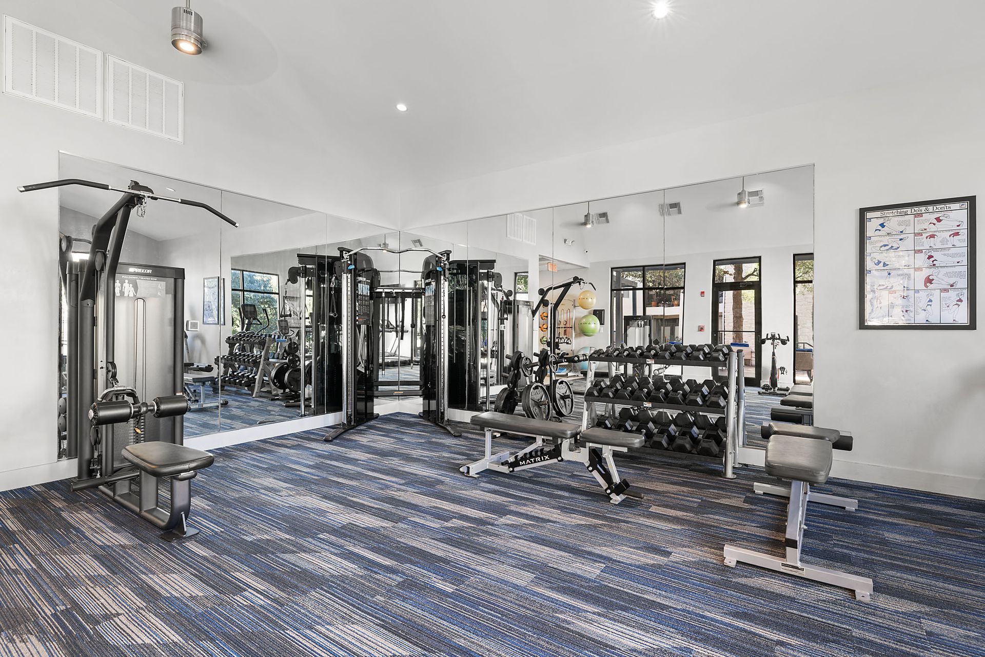 Bright gym interior with cable machines, mirrors, dumbbells, and blue patterned carpet at Marquis at Great Hills in Austin, TX.