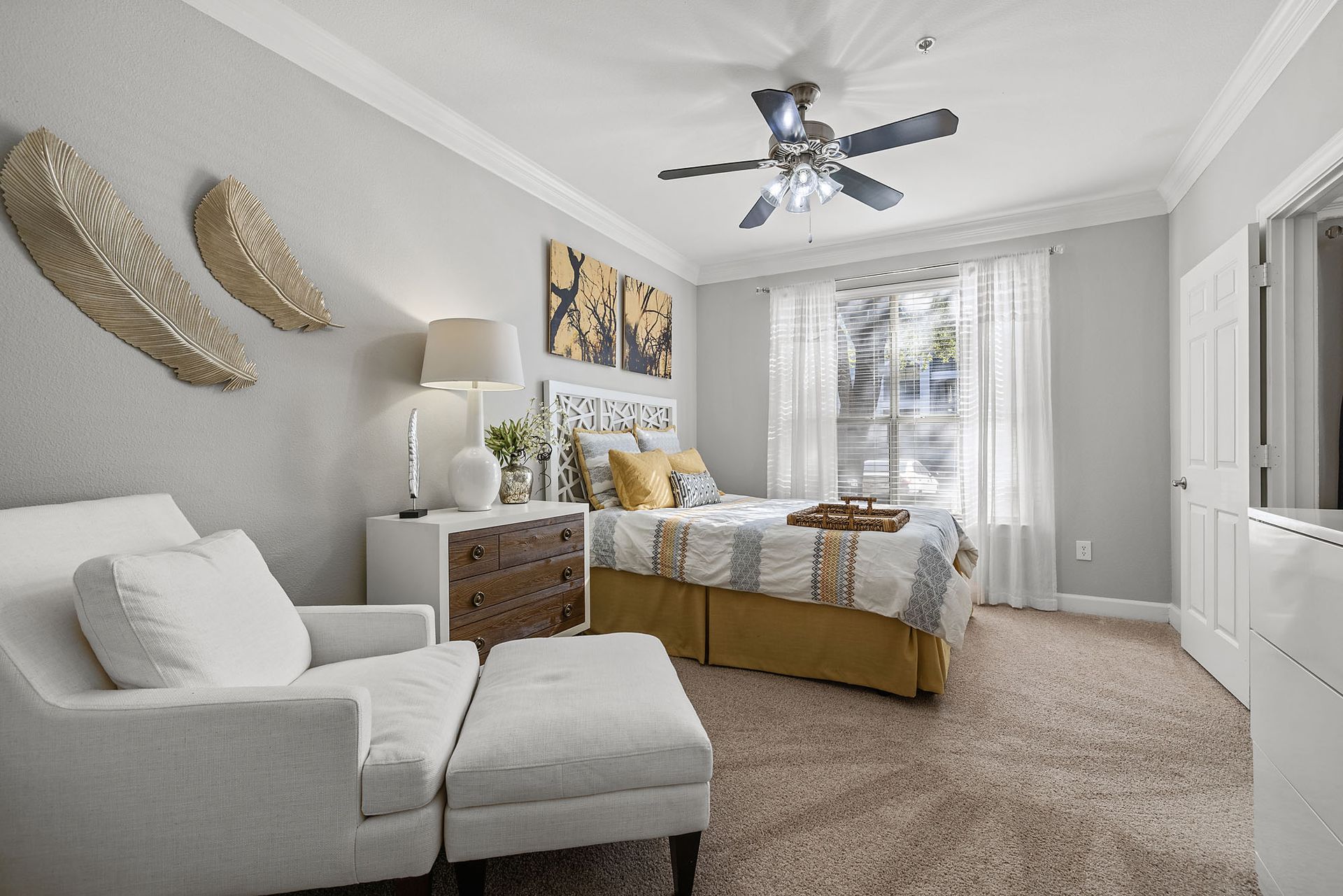 Bright bedroom with white chaise lounge, beige bed, patterned decor, and a ceiling fan near a window at Marquis at Great Hills in Austin, TX.