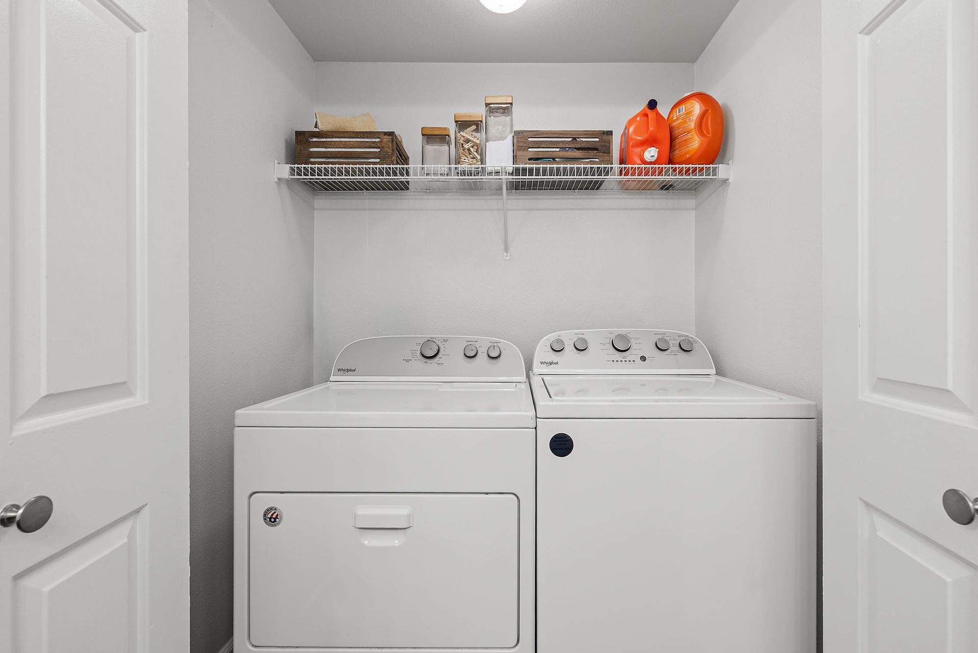 Small laundry room with white washer and dryer, wire shelf, and orange detergent bottles at Marquis at Great Hills in Austin, TX.