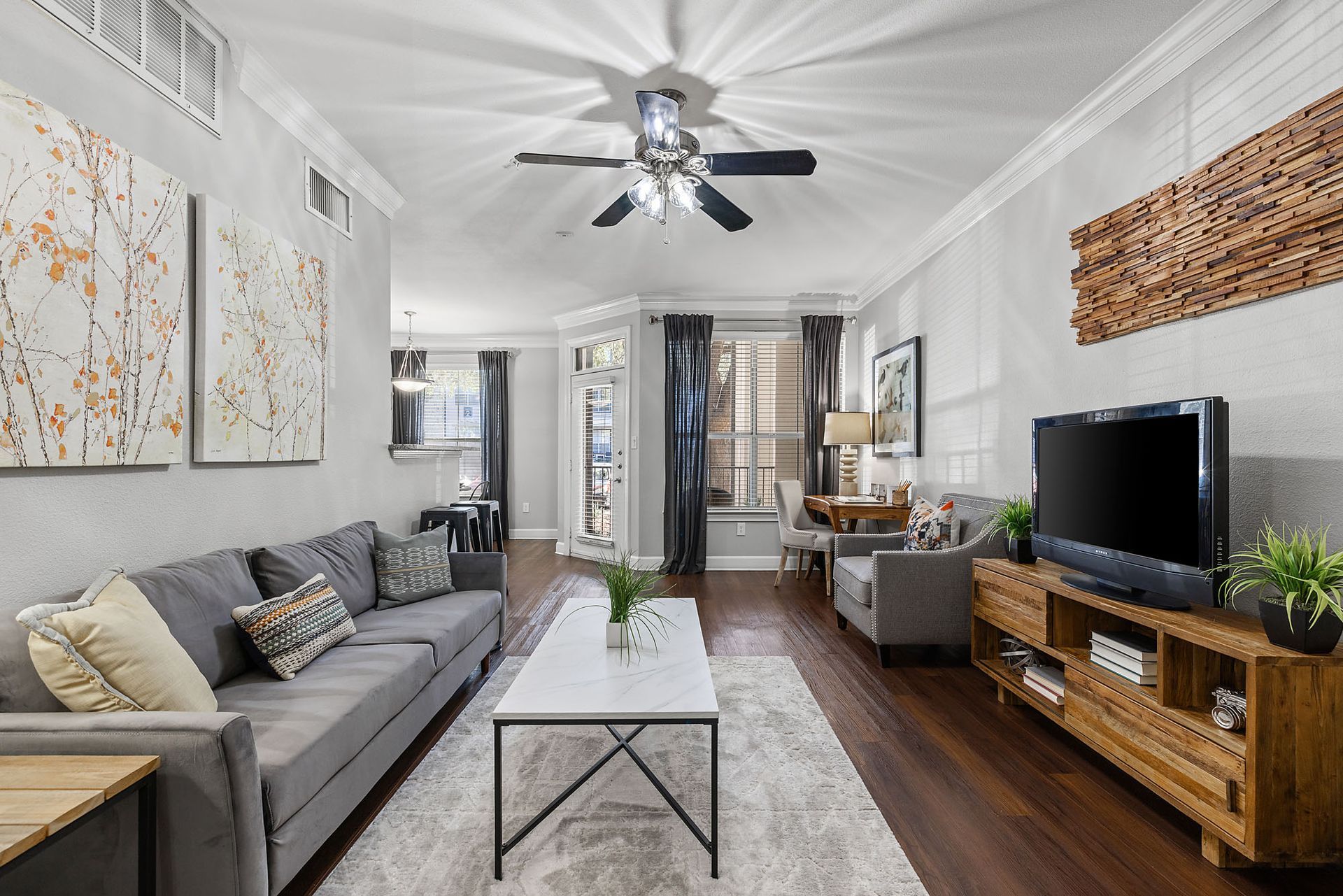 Bright living room with gray sofa, TV, wood floor, ceiling fan, and white walls at Marquis at Great Hills in Austin, TX.