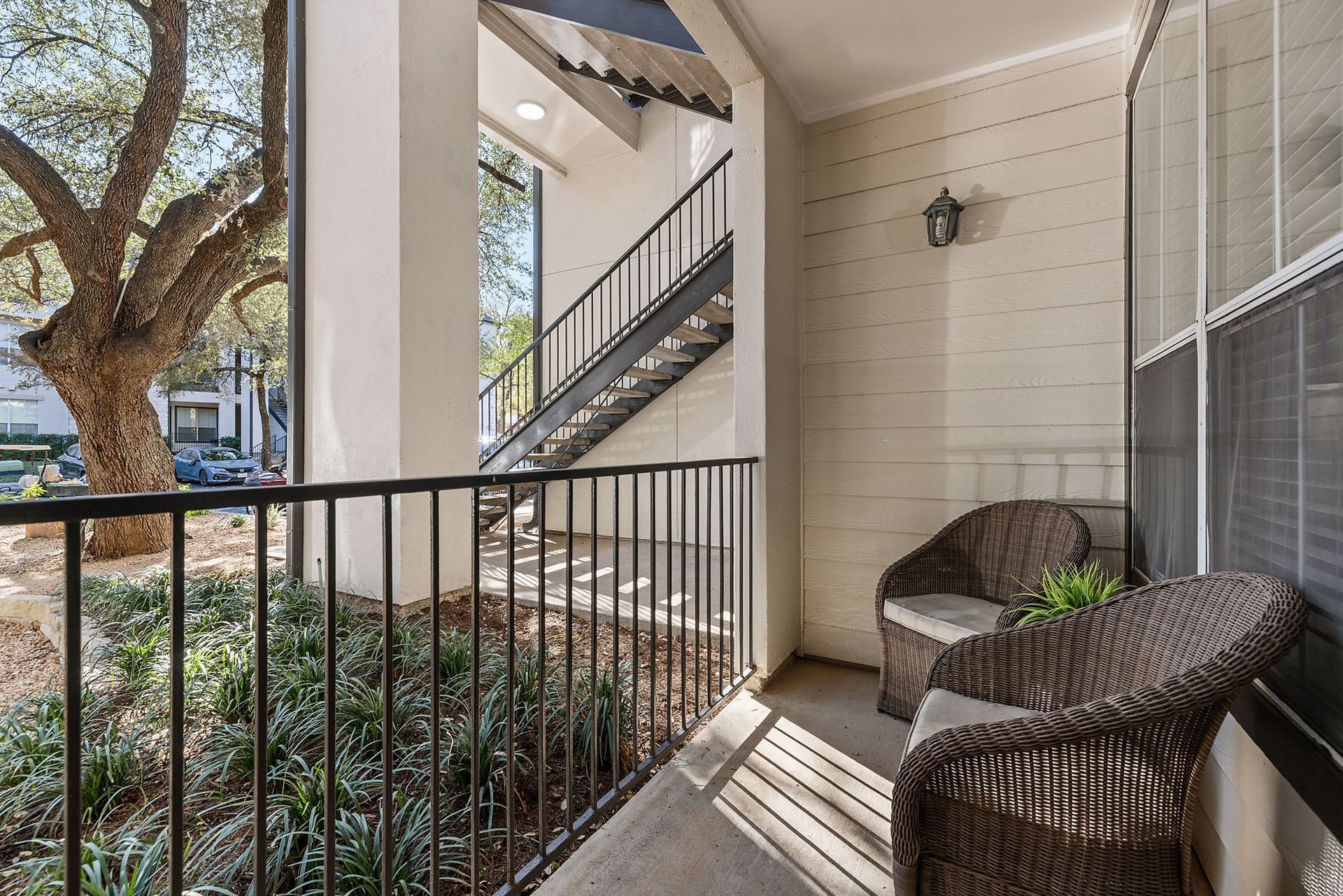 Apartment balcony with wicker chairs, potted plant, and stairs beside a railing in sunlight at Marquis at Great Hills in Austin, TX.