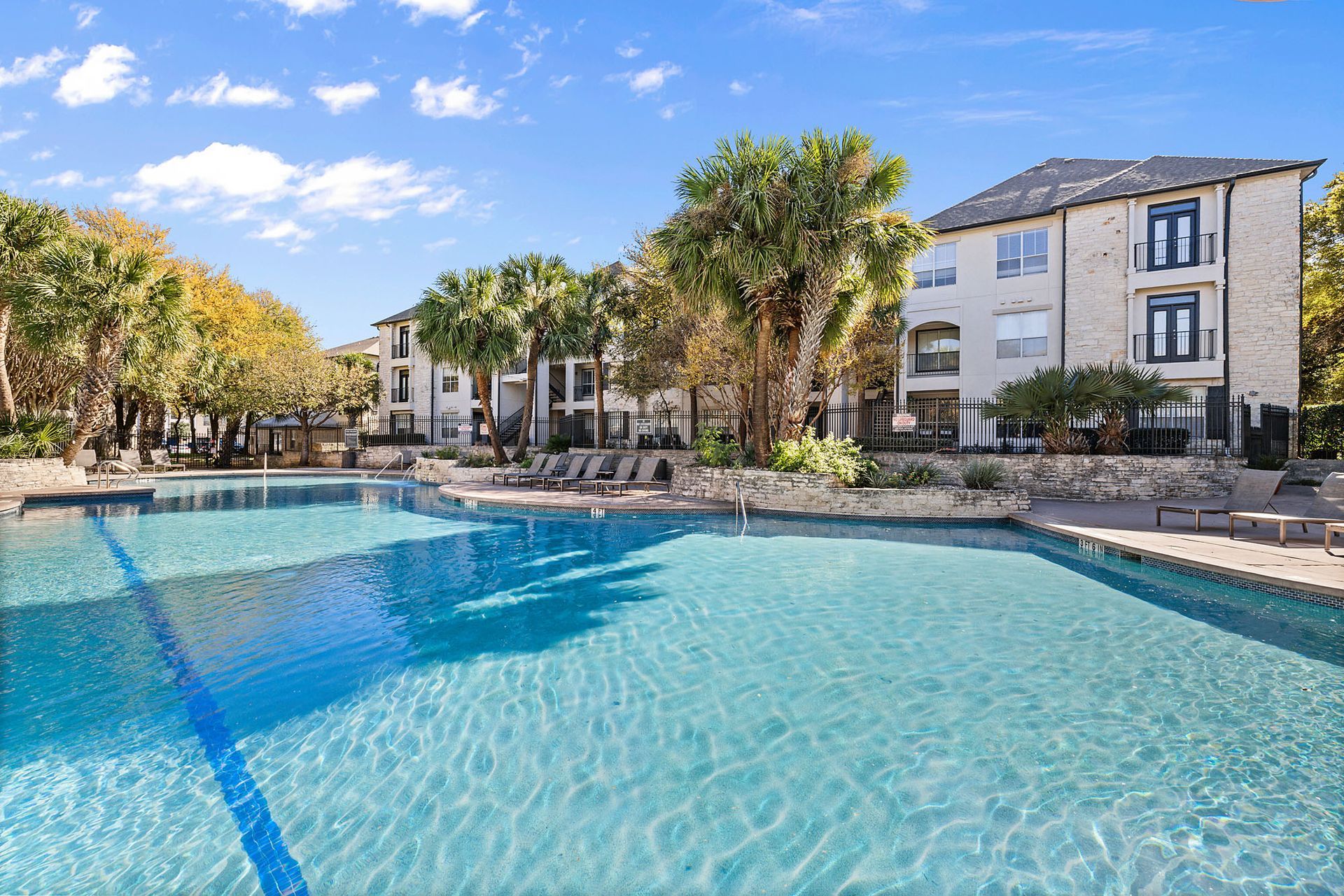 Sunny resort pool surrounded by palm trees and white buildings under a blue sky at Marquis at Great Hills in Austin, TX.