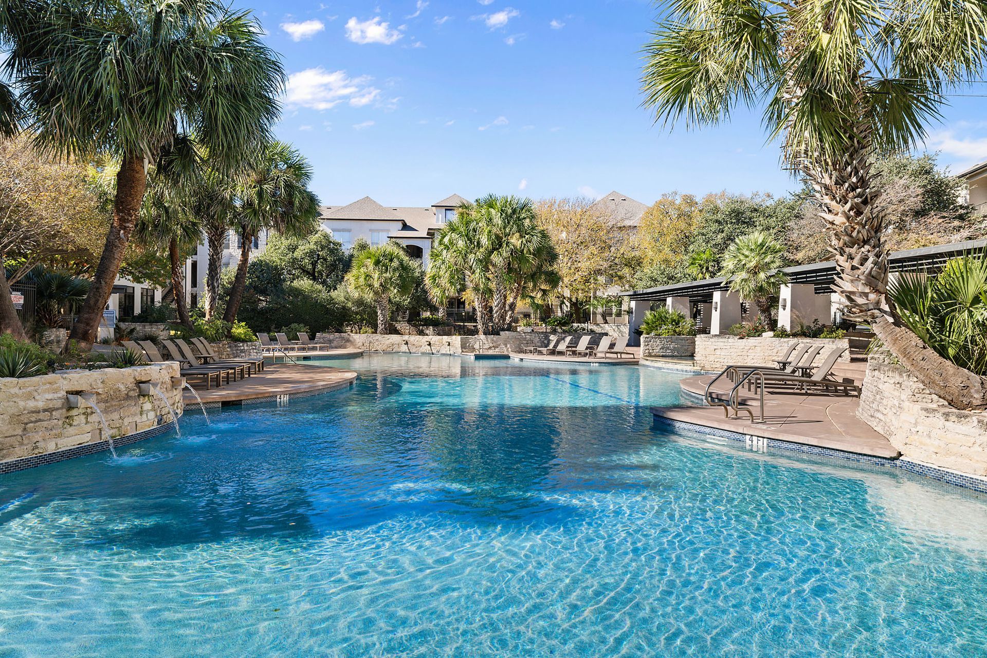 Outdoor resort swimming pool with clear blue water, rocks, trees, and lounge area in the background at Marquis at Great Hills in Austin, TX.