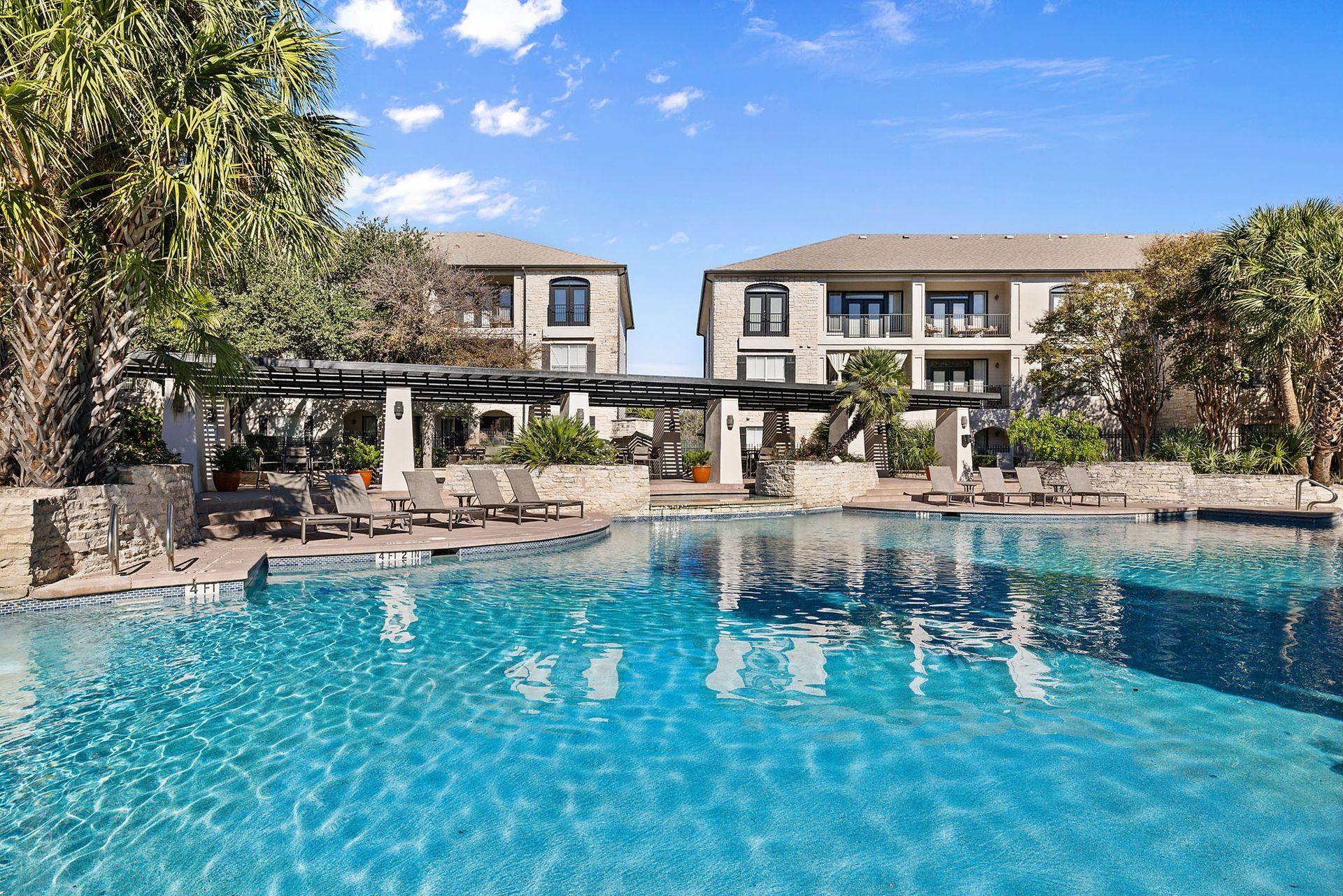 Resort swimming pool with blue water, stone deck, lounge chairs, and palm trees under a clear sky at Marquis at Great Hills in Austin, TX.