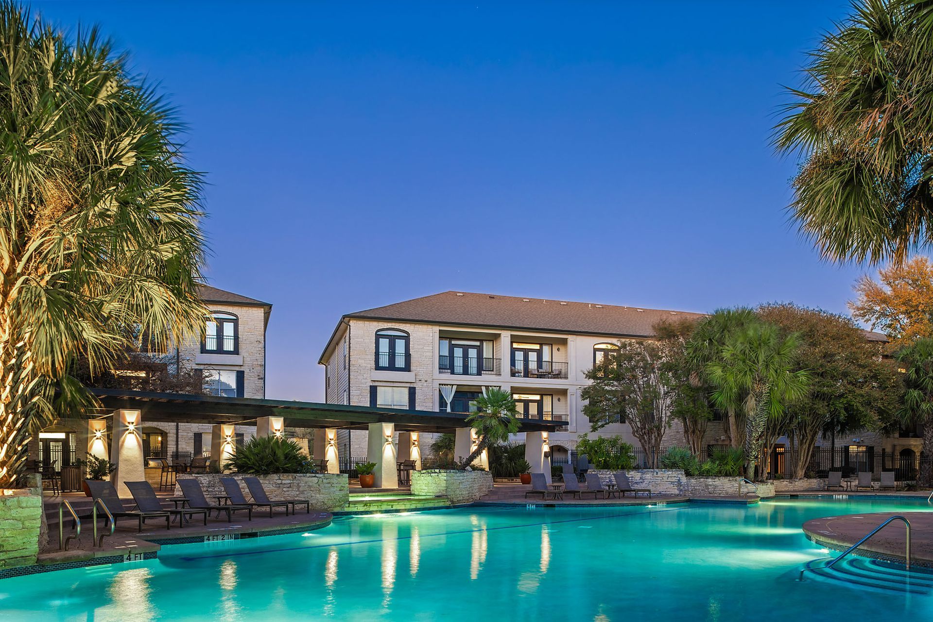 Luxury resort with lit pool, palm trees, and a two-story building at dusk at Marquis at Great Hills in Austin, TX.