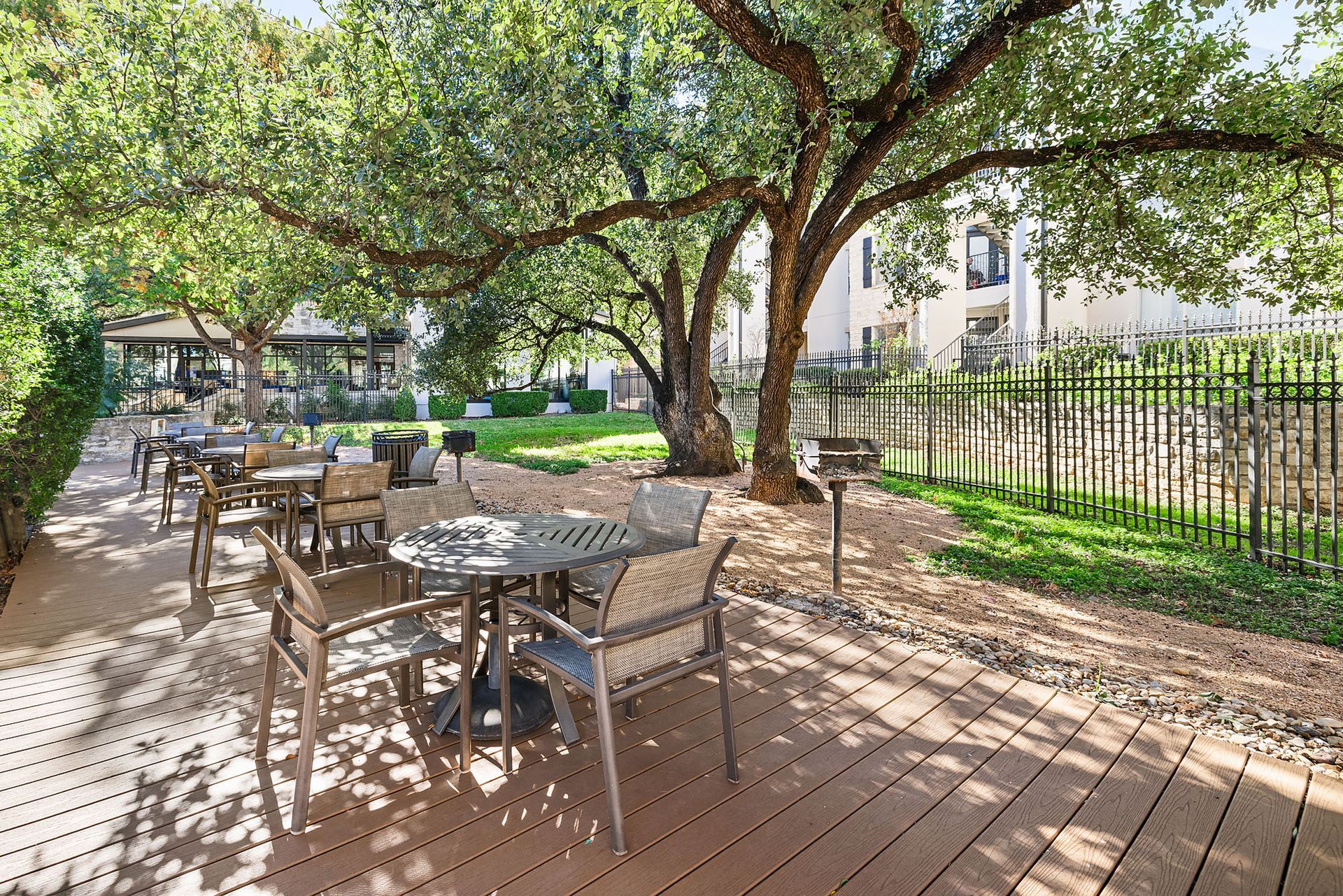 Shaded outdoor café patio with empty tables and chairs under leafy trees beside a fence and building at Marquis at Great Hills in Austin, TX.